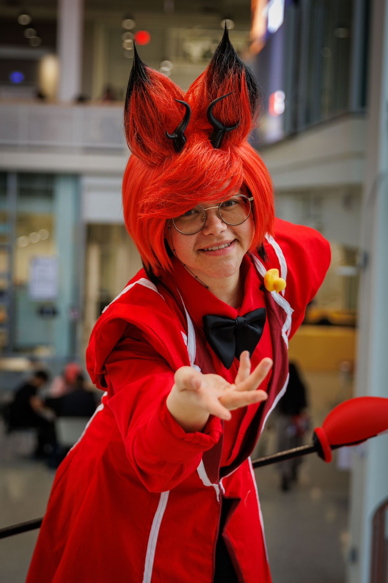Person cosplaying as Alastor from Hazbin Hotel in red outfit, fox ears wig, bow tie, reaching toward camera.
