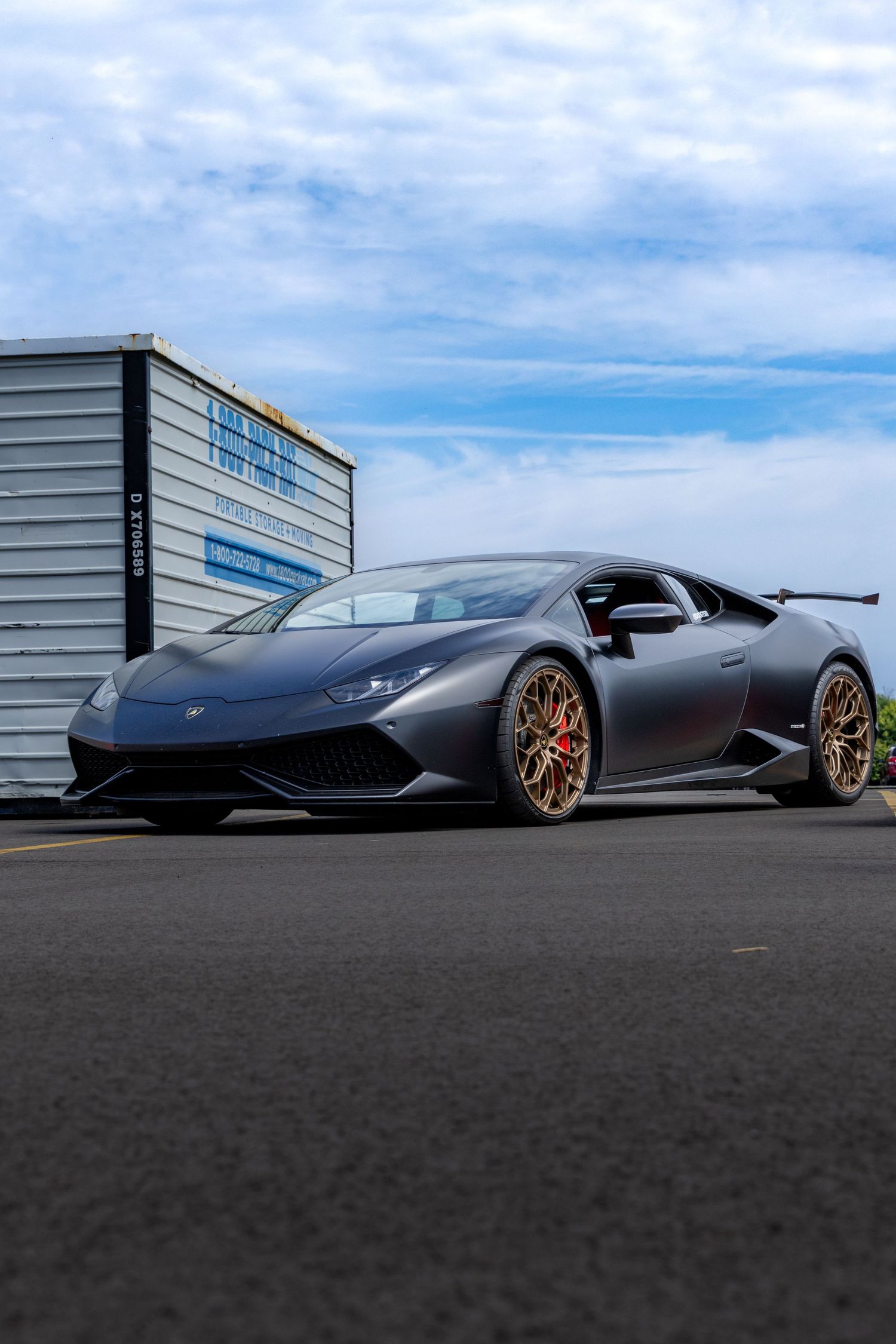 Sleek matte black Lamborghini sports car with custom bronze wheels parked on asphalt against industrial building backdrop.