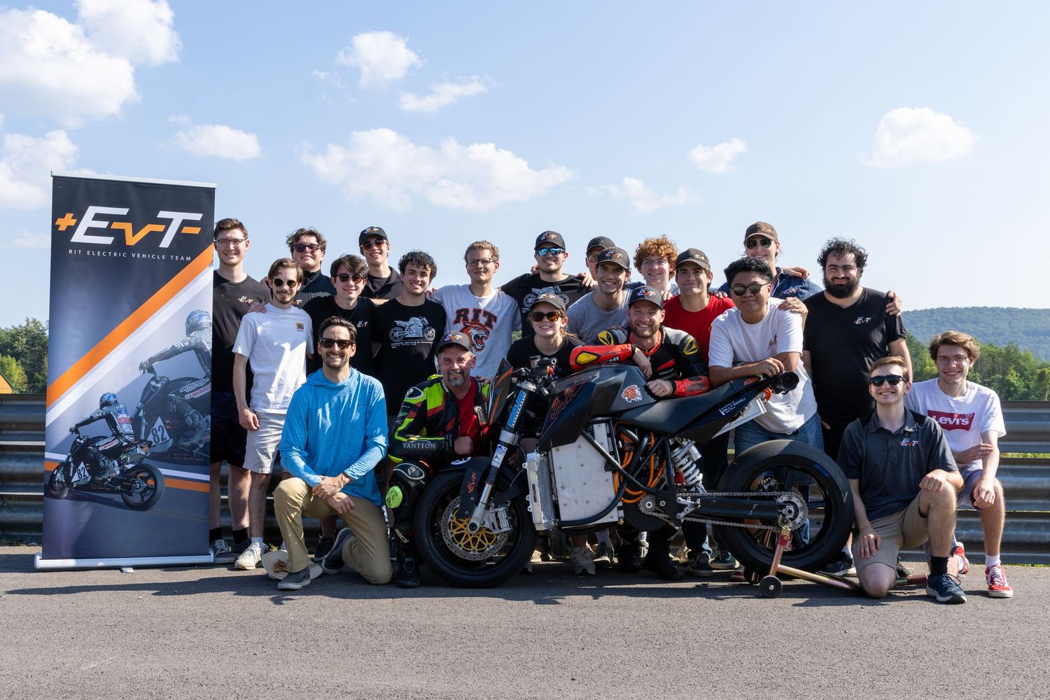 A group of people sitting together outdoors with motorcycles parked nearby during a sunny day.