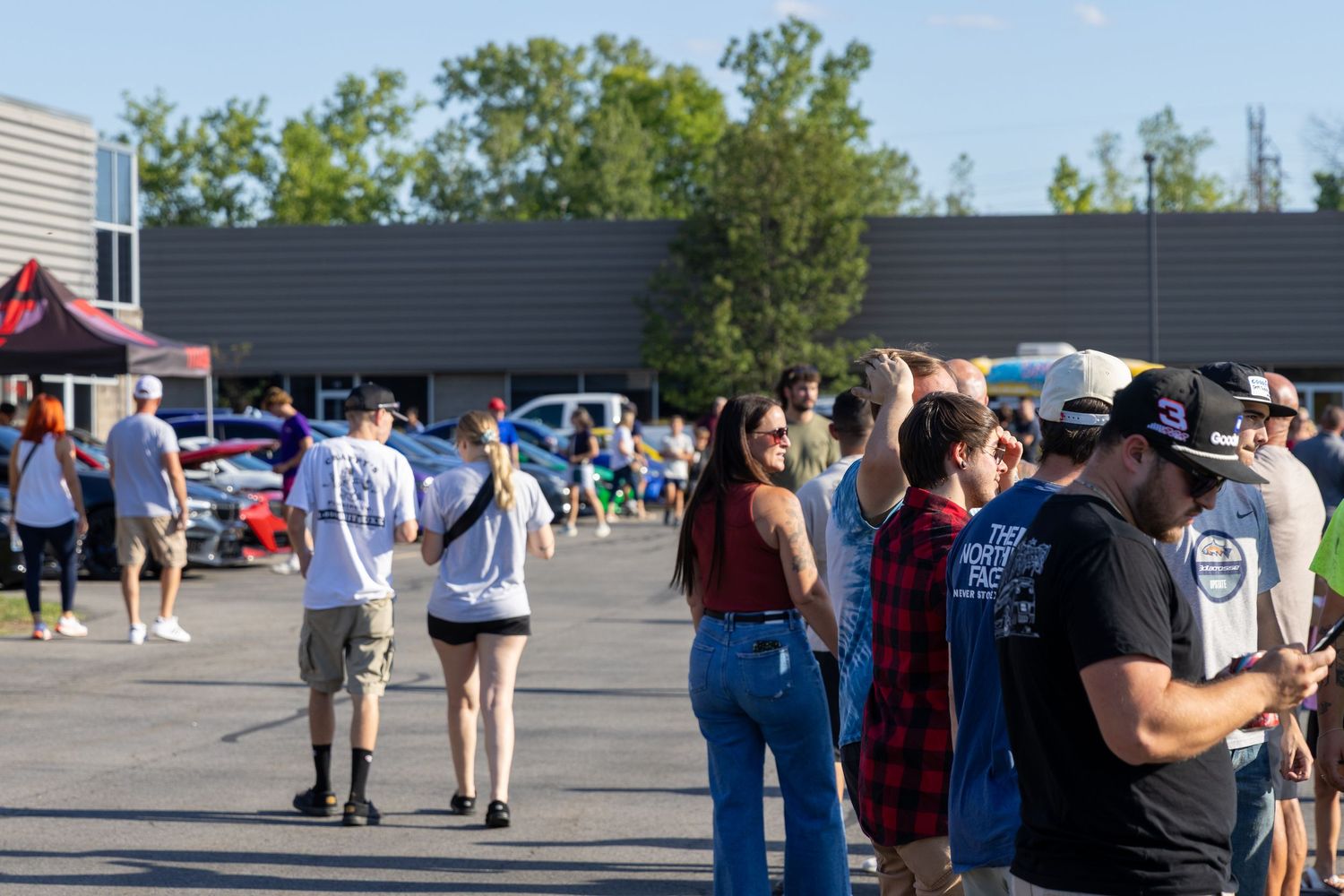 A long line of people wait outside a building on a sunny day in a commercial parking lot.