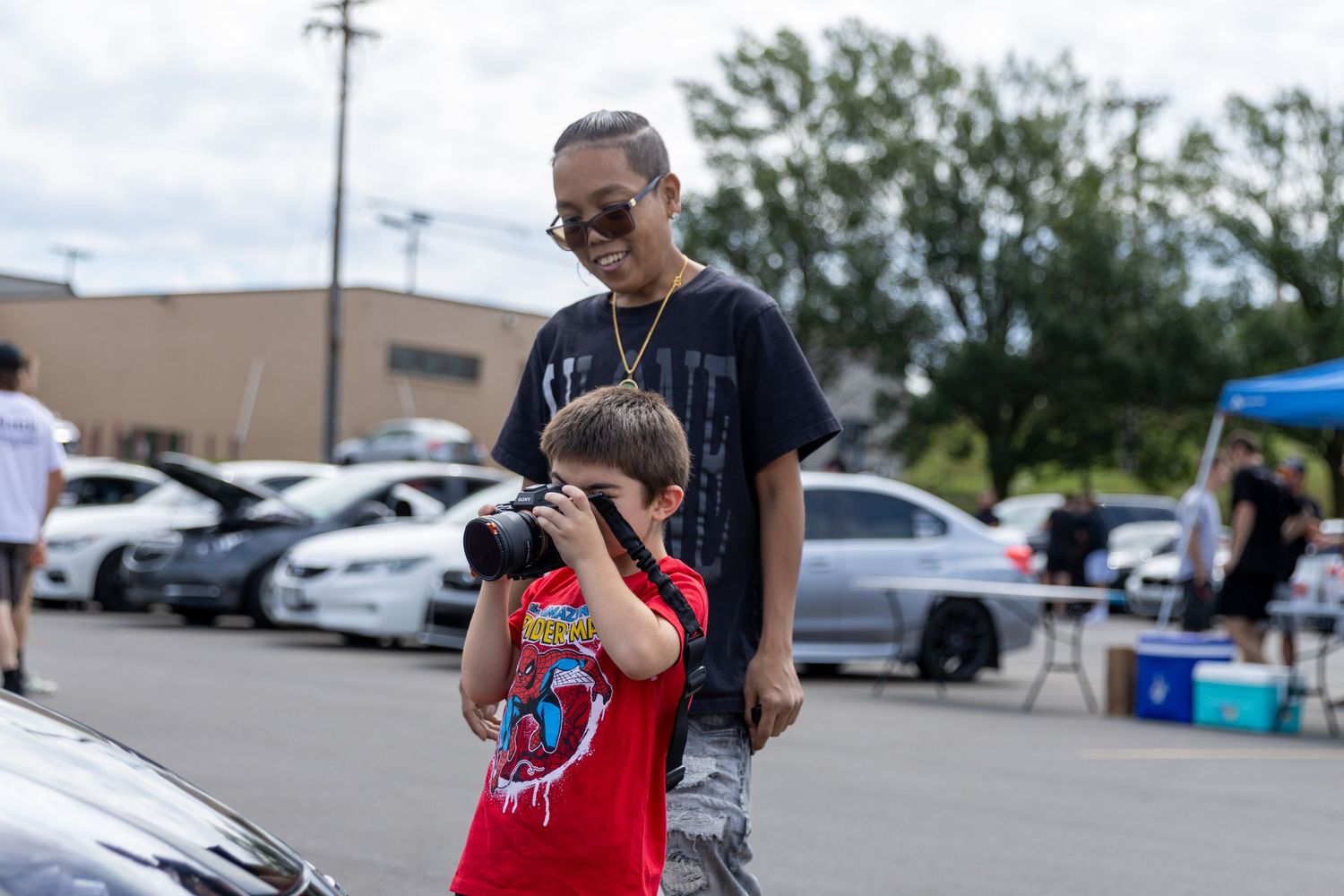 People in a parking lot browse items at an outdoor shopping event on a sunny day.