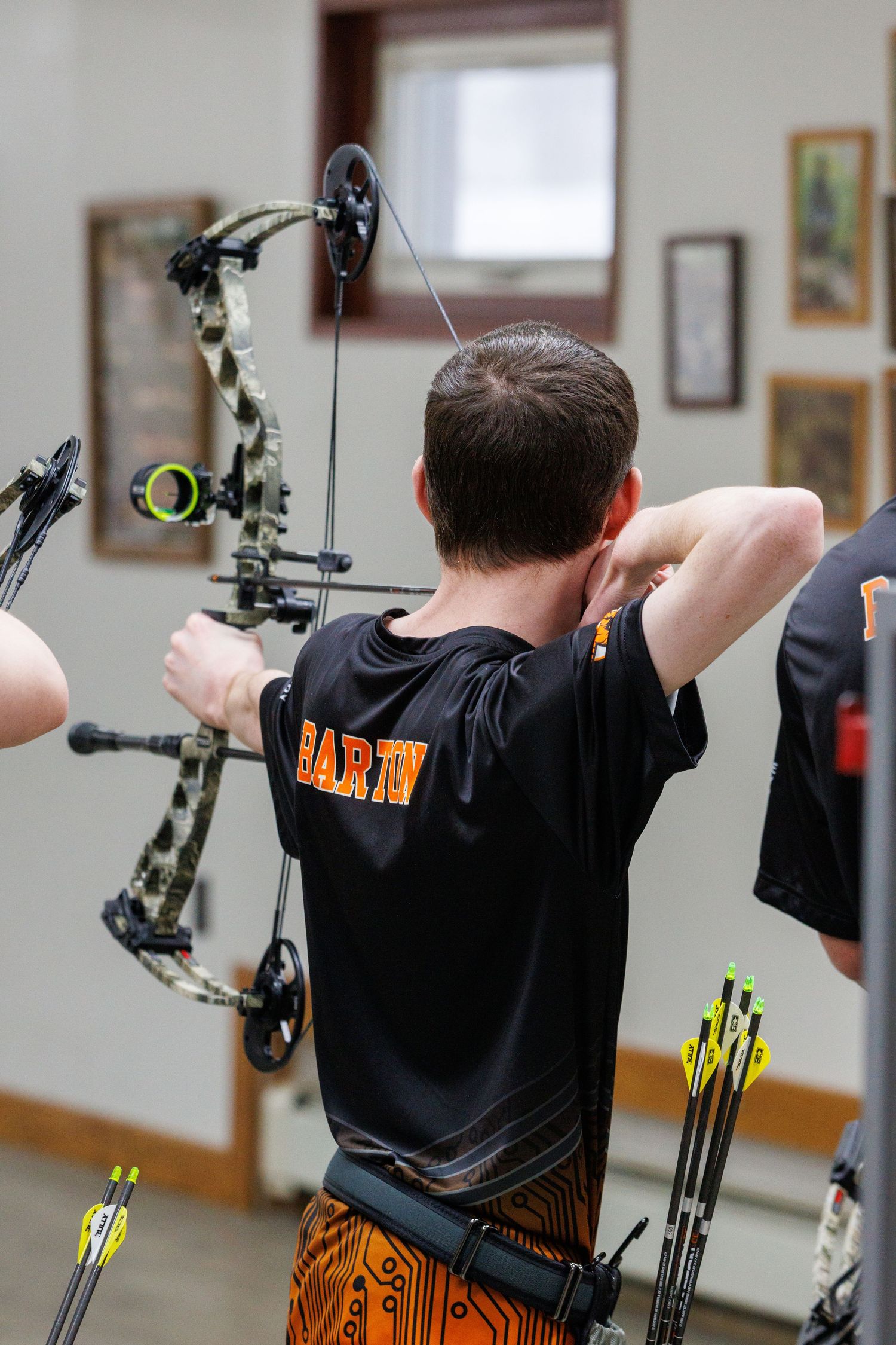 Archer in a black Barton jersey draws a compound bow indoors during an archery competition.