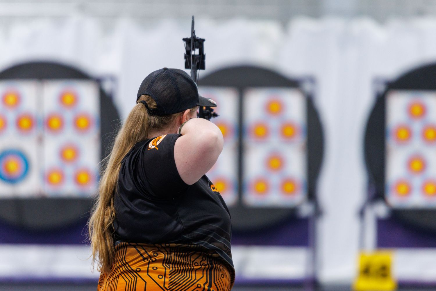 Female archer with long hair aims a compound bow at circular archery targets outdoors.