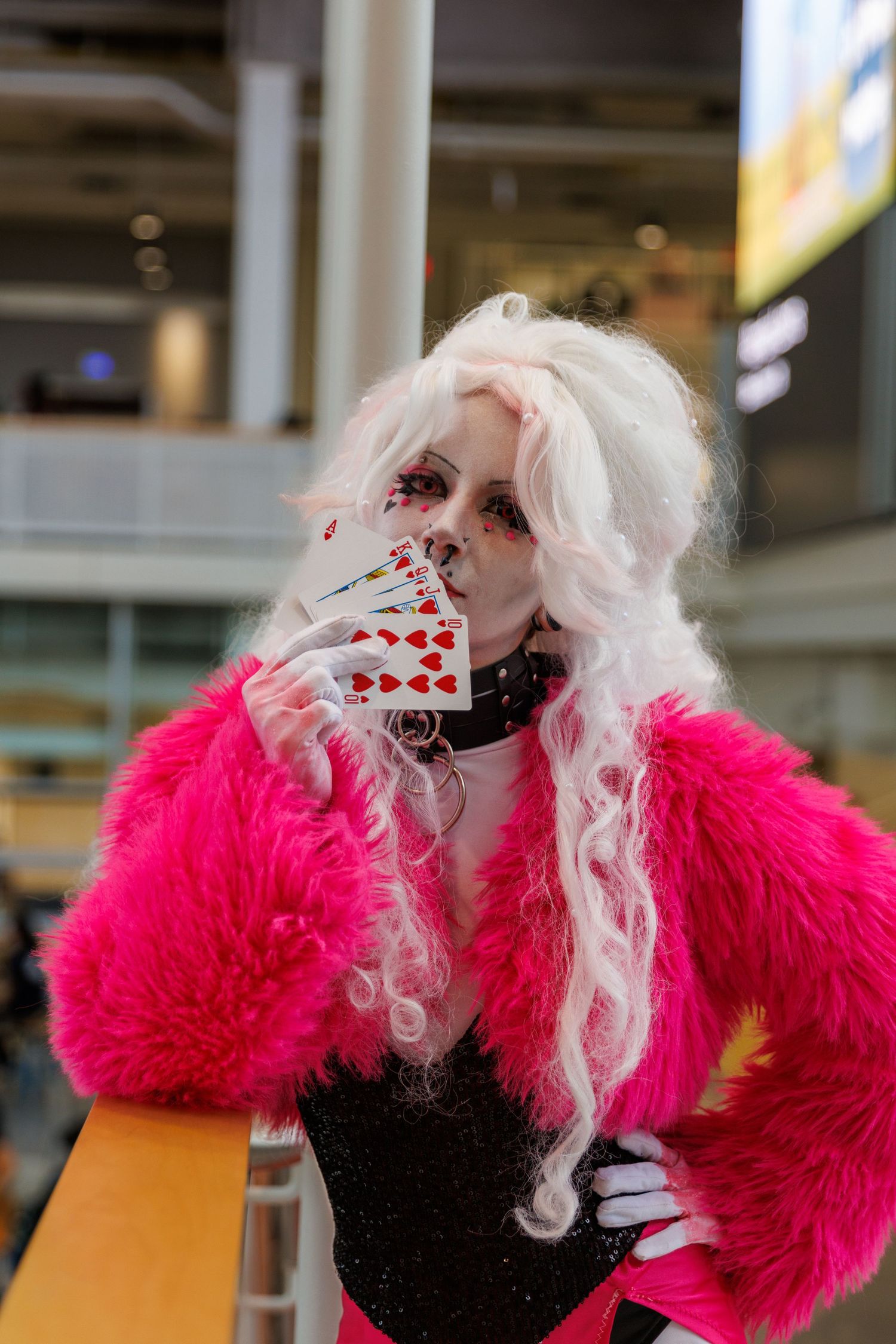 Cosplayer in pink fur coat and white wig holds playing cards over face with dramatic makeup at convention.