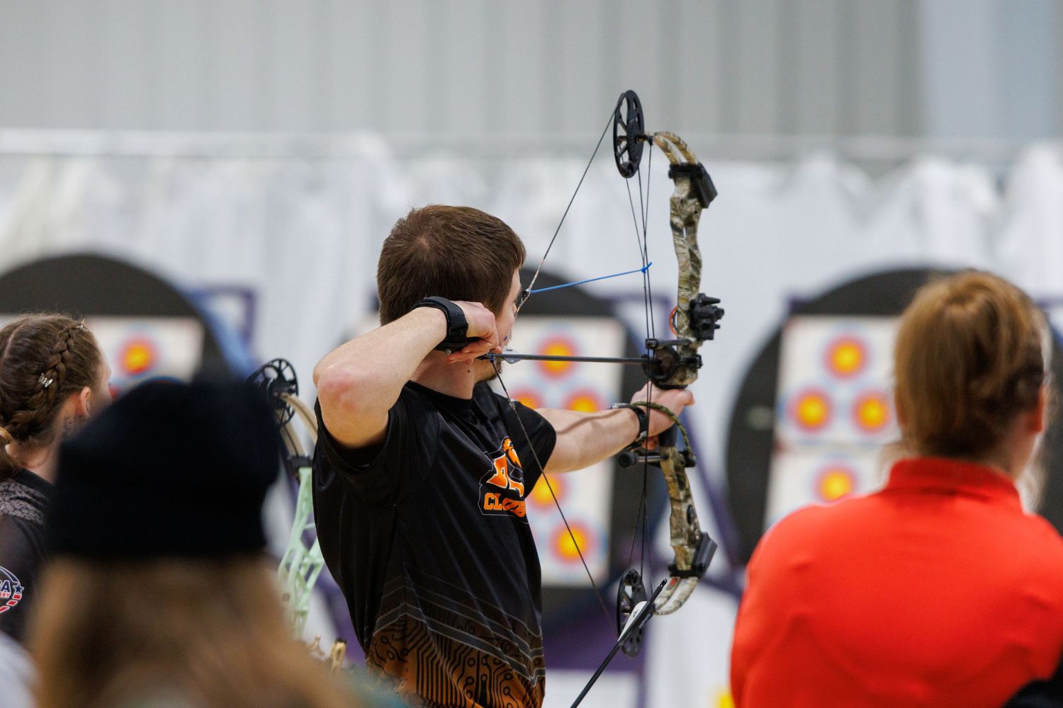 Young archer aiming compound bow at targets during indoor archery competition.