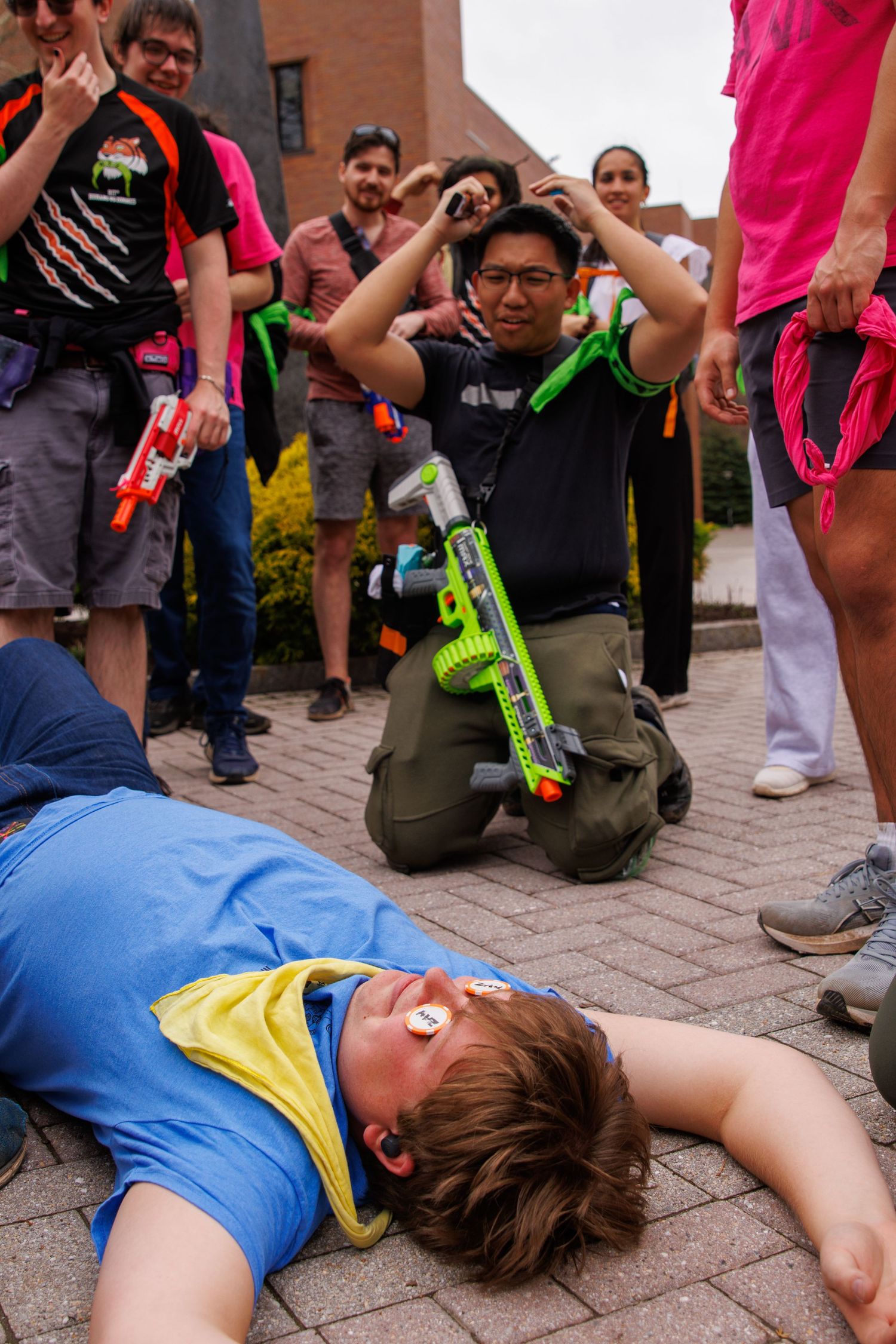 Group of people playing Nerf gun battle outdoors, with one player lying on ground defeated and another kneeling with green blaster.