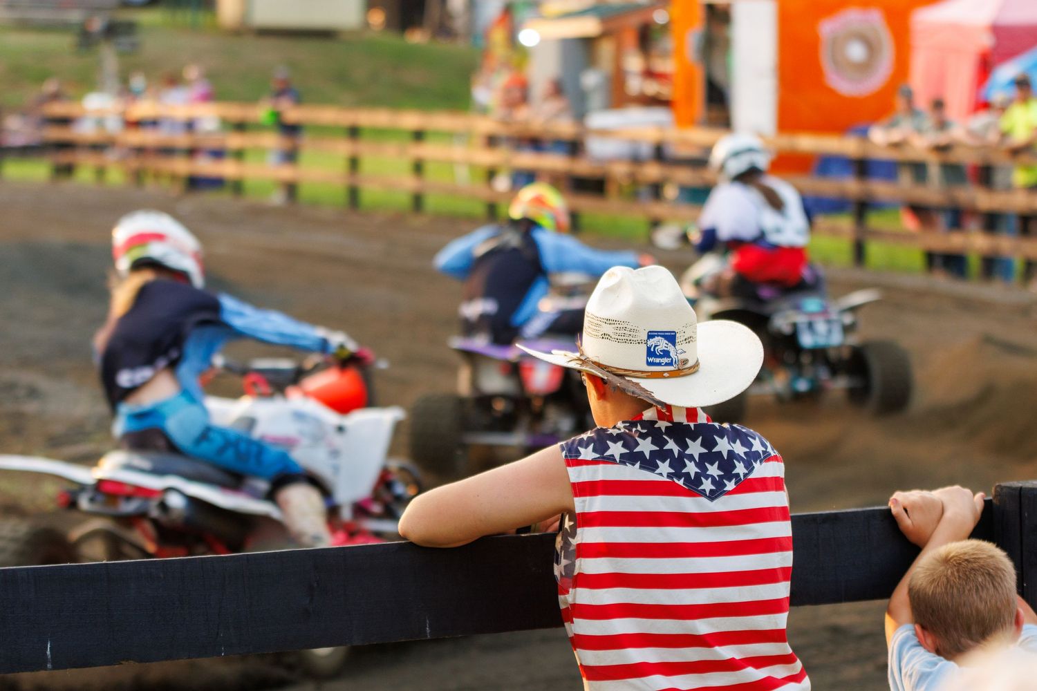 Person in American flag tank top and cowboy hat watches ATV riders race at an outdoor motocross event.