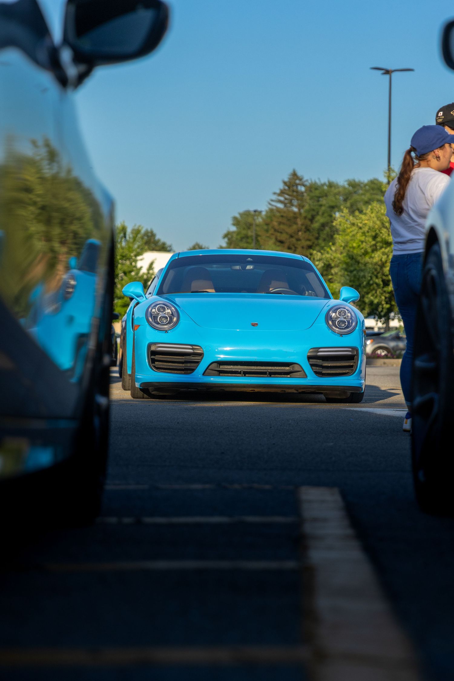 Light blue Porsche 911 sports car viewed between other vehicles in a parking lot.