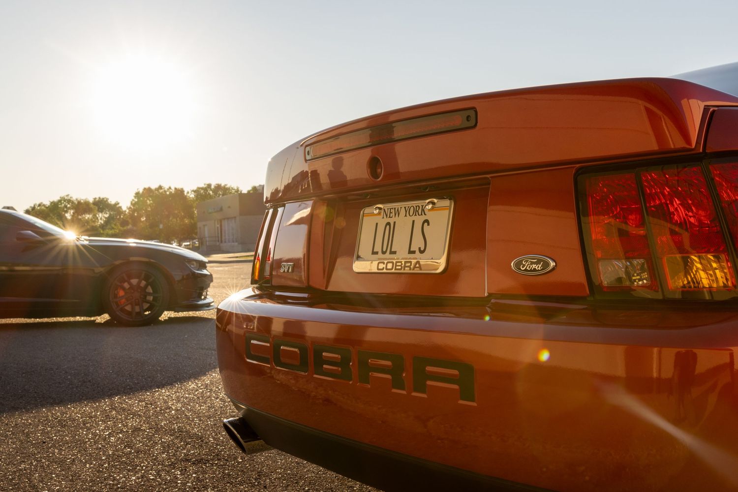 Orange Ford Mustang Cobra rear end with tail lights glowing at sunset in a parking lot.