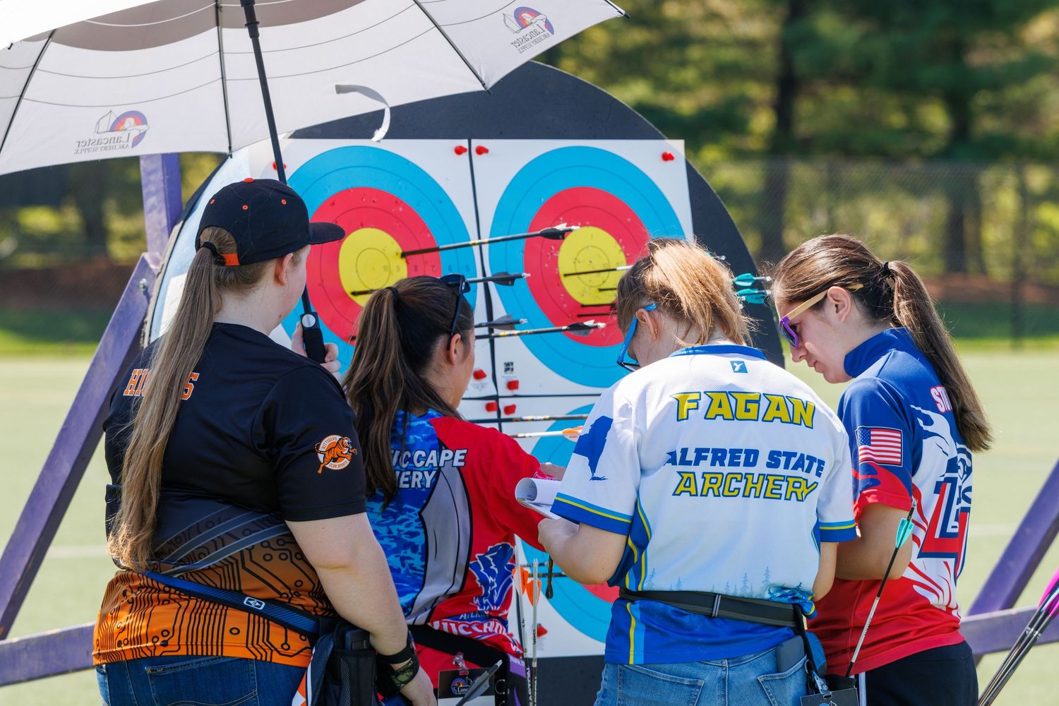 Archery team members huddle around a target during an outdoor competition, wearing Alfred State Archery uniforms.