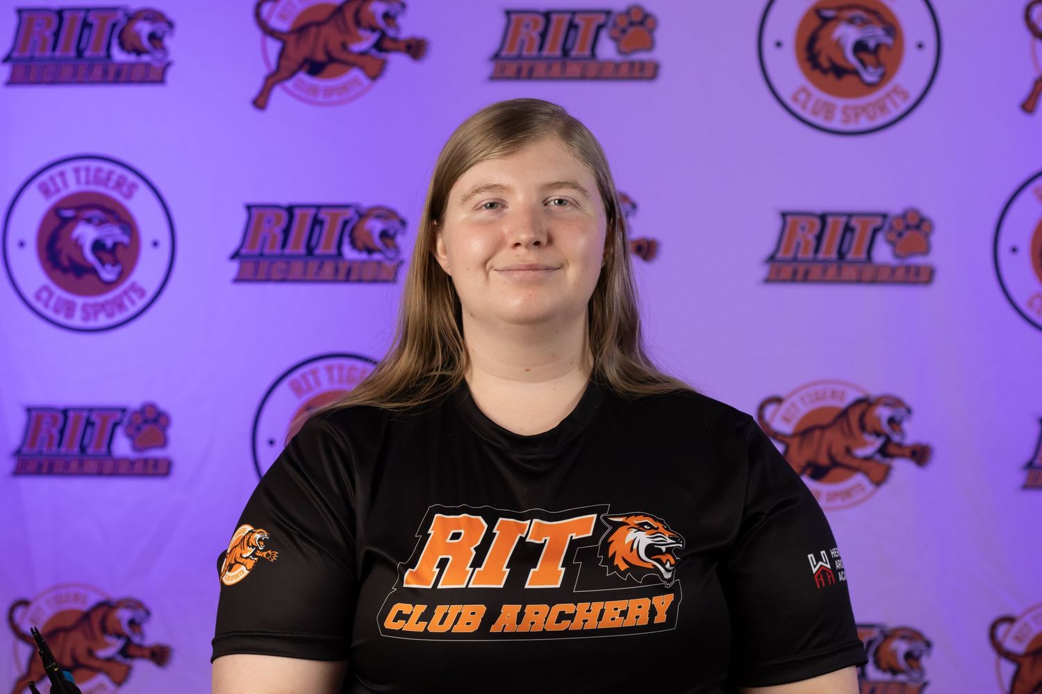 Student athlete in RIT Club Hockey shirt poses against purple backdrop with tiger logos.
