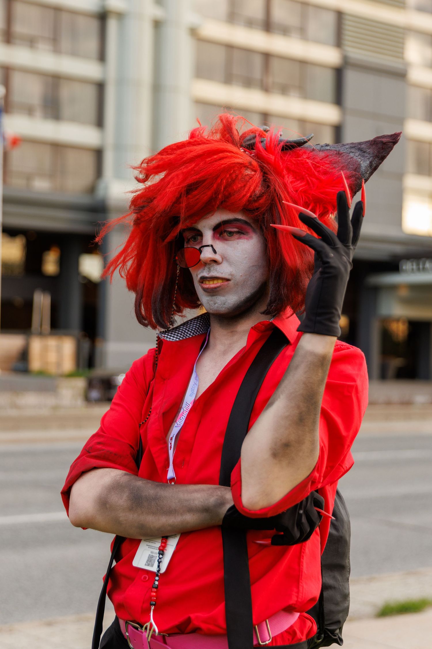 Person in red wig and gray face paint cosplaying a demon character with claws at an outdoor event.