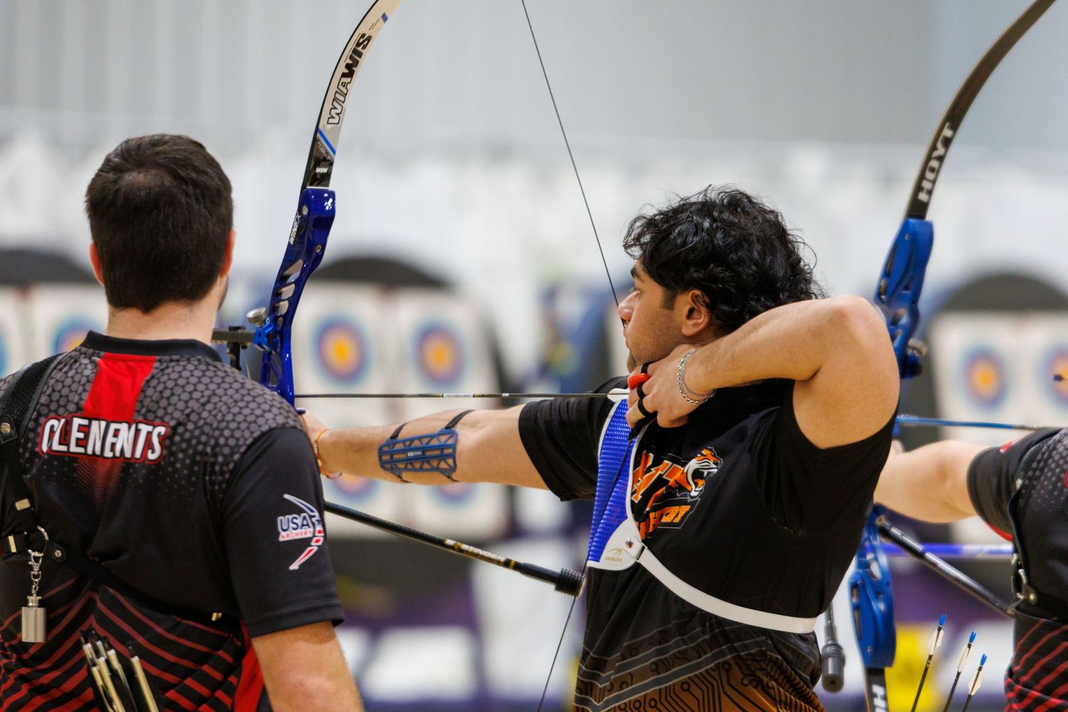 Two male archers drawing recurve bows at an indoor archery range with targets in background.