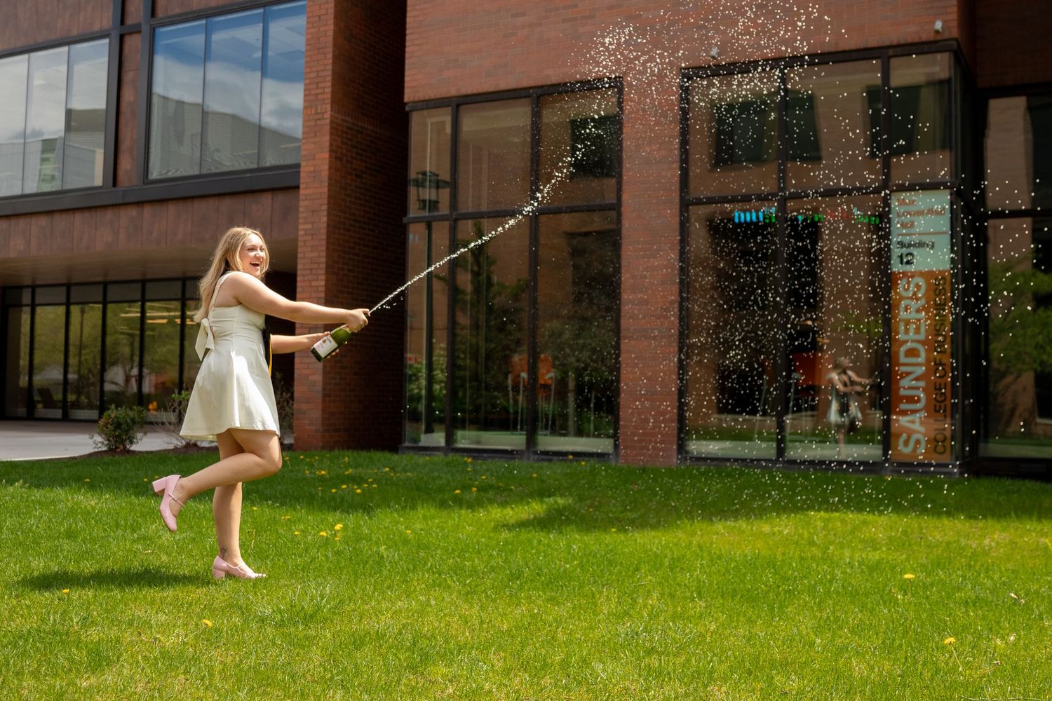 Person in a white dress uses a sprinkler on a sunny day outside a brick building.