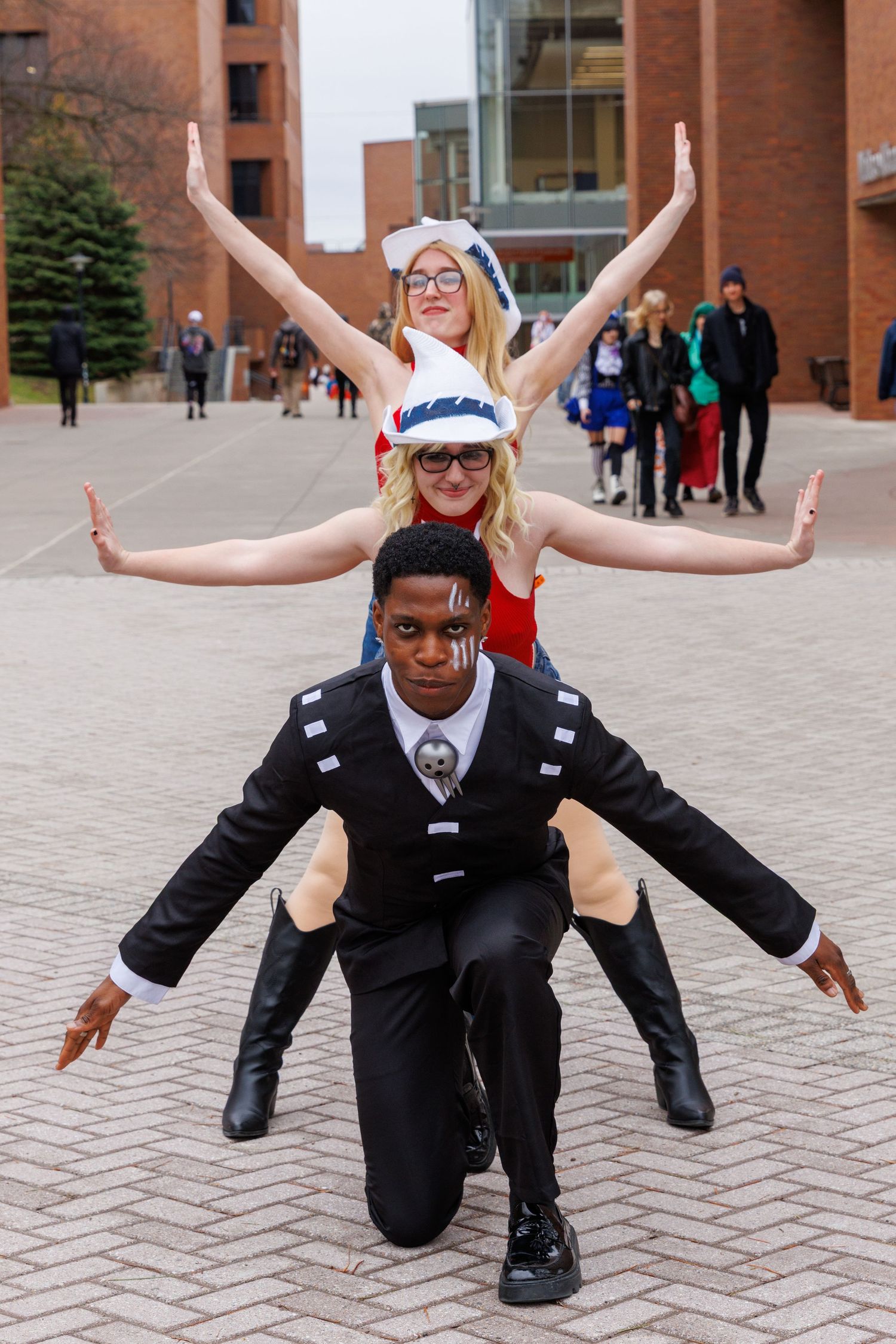 Three cosplayers pose dramatically outdoors, with two forming a human tower with arms spread wide.
