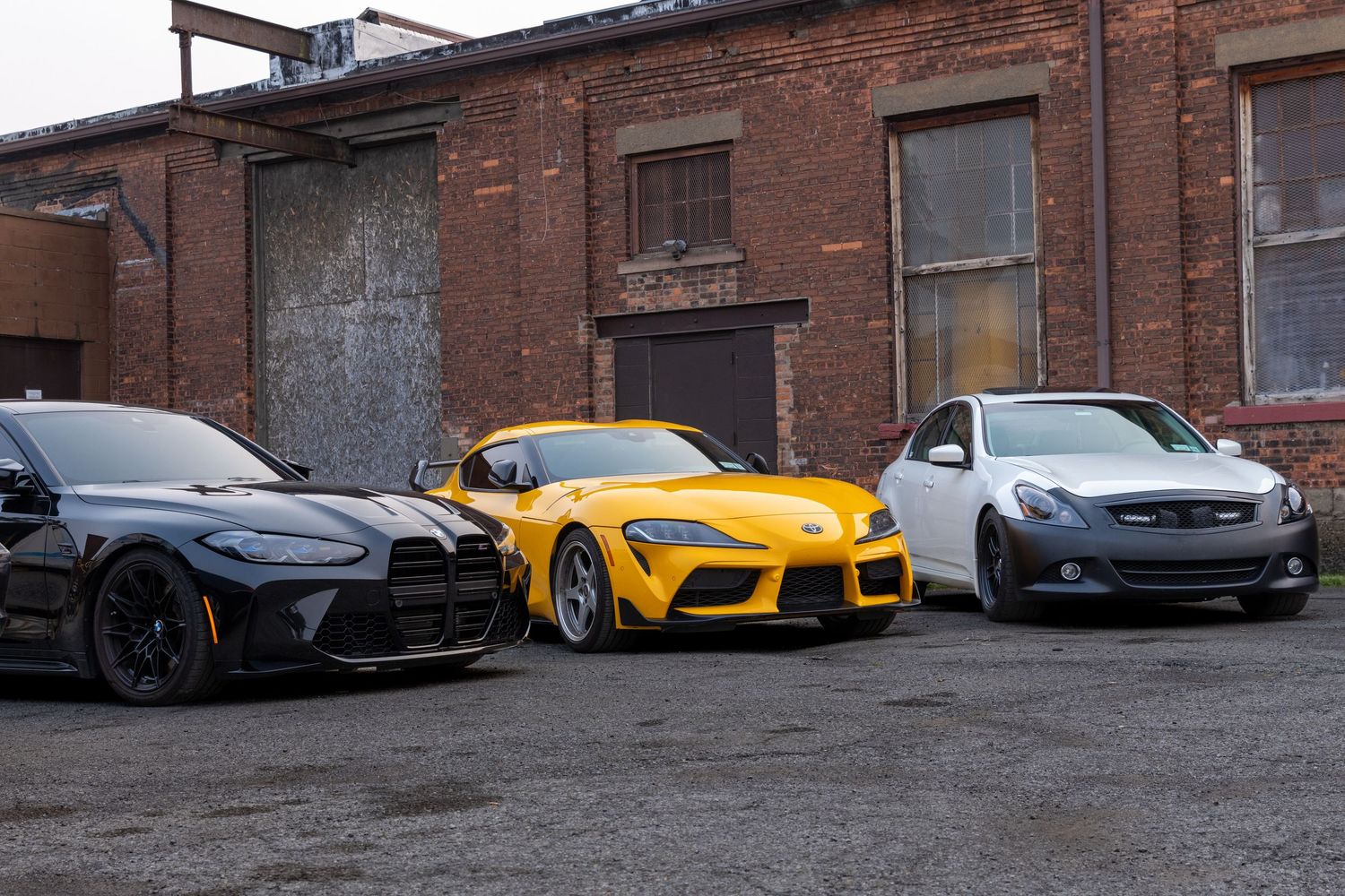 Row of luxury sports cars including a black Porsche, yellow Toyota Supra, and dark sedan parked near brick buildings.