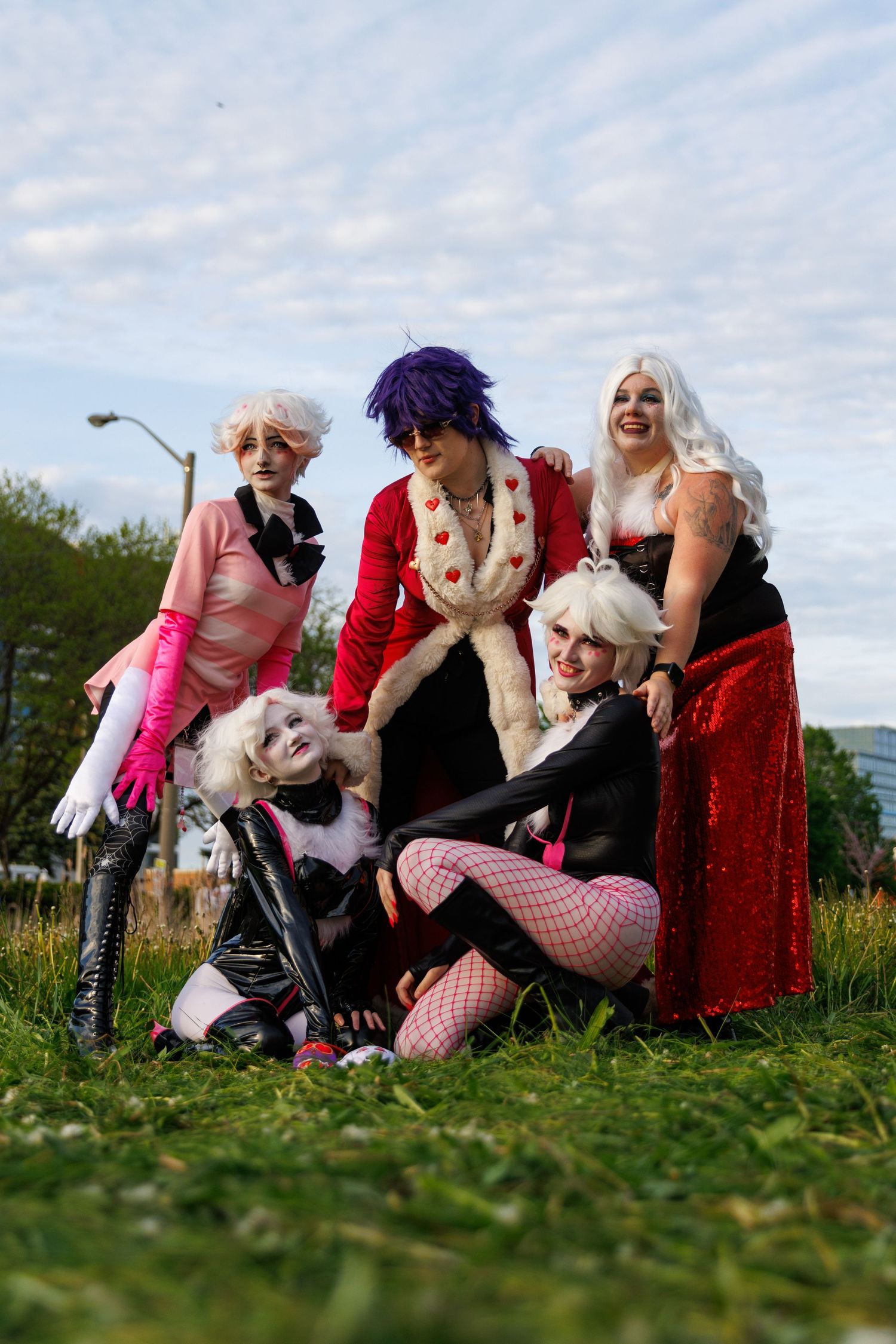 Group of five cosplayers in elegant villain-inspired costumes posing together on grass at outdoor event.