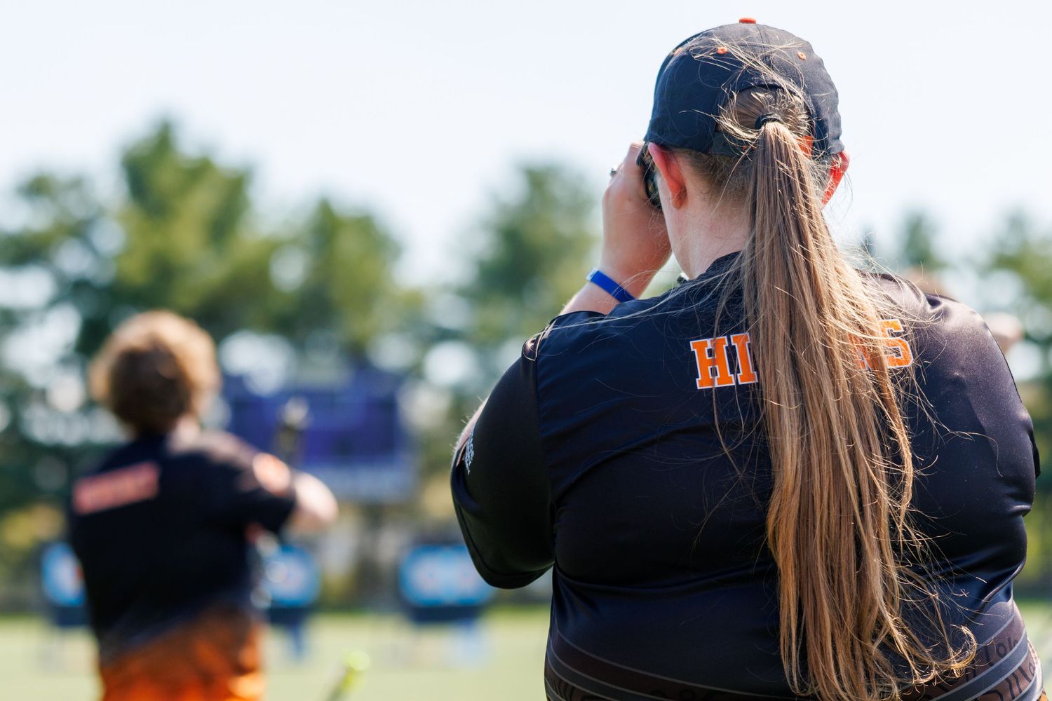 A female archer in an orange and navy uniform uses binoculars to observe targets at an outdoor archery event.