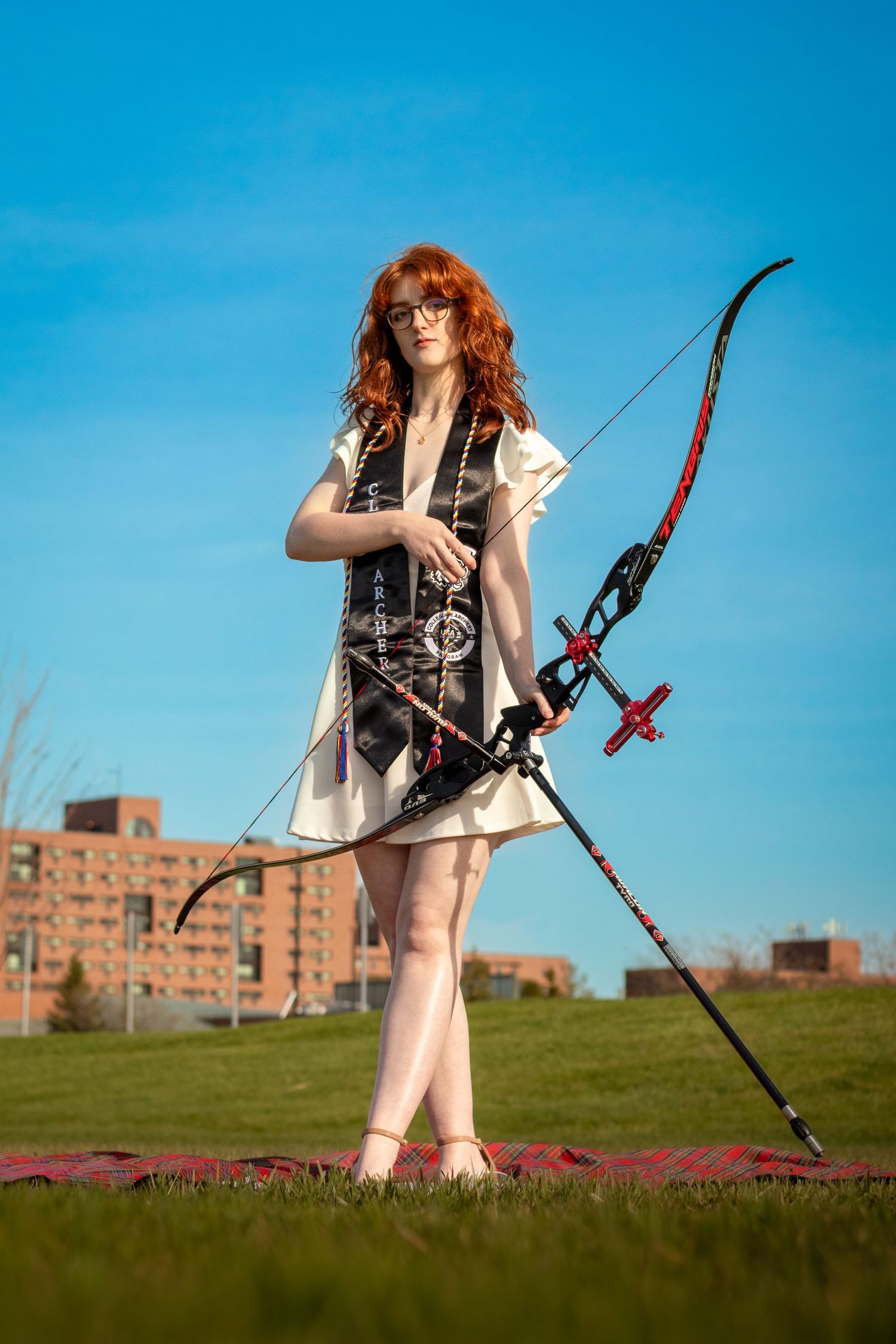 Archer in a white dress poses with a modern compound bow against a bright blue sky background.