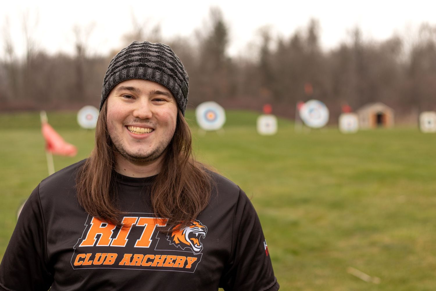 Person wearing knit cap and archery club shirt smiles at outdoor archery range.