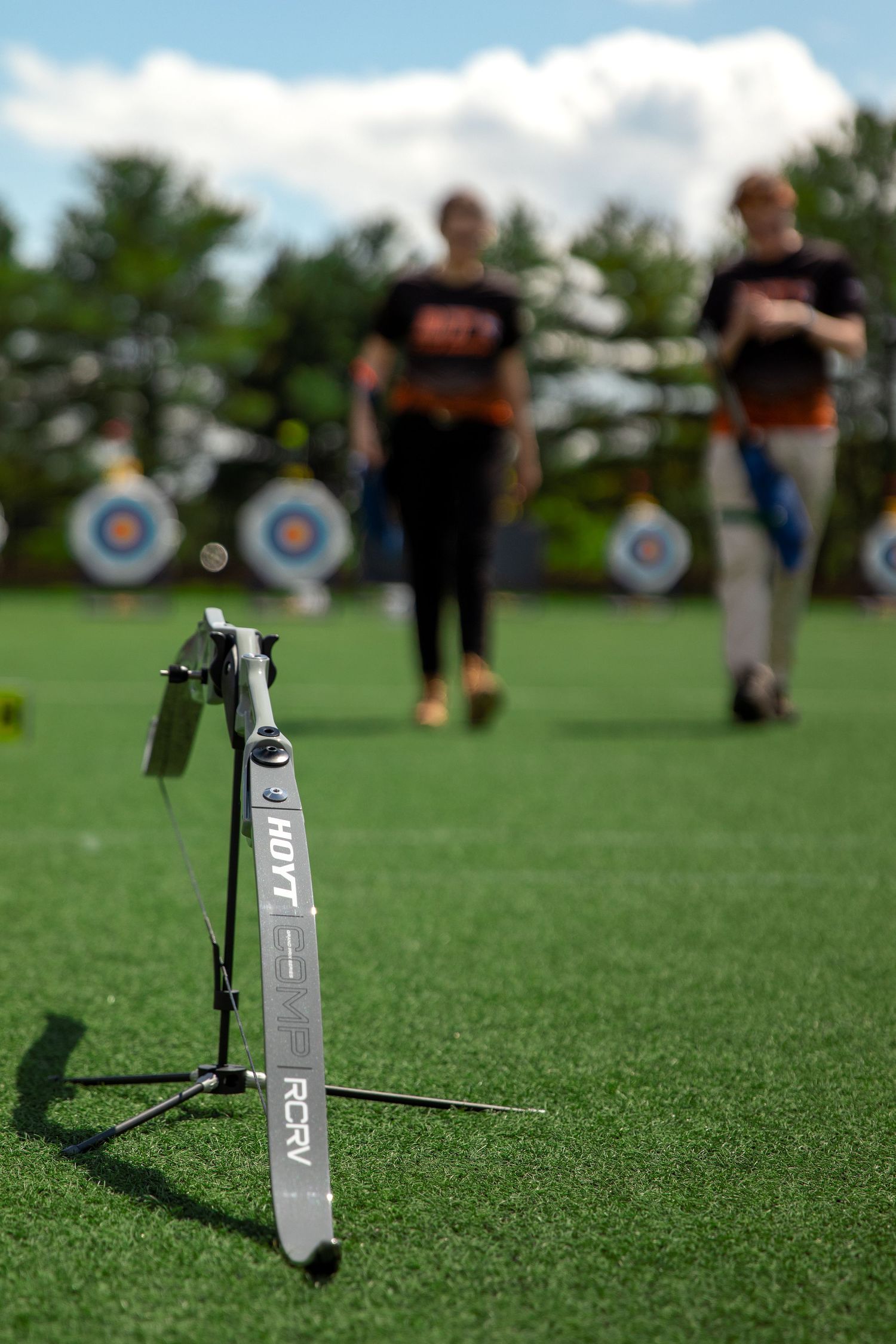 Archery bow on stand with blurred figures and targets in background on sports field.