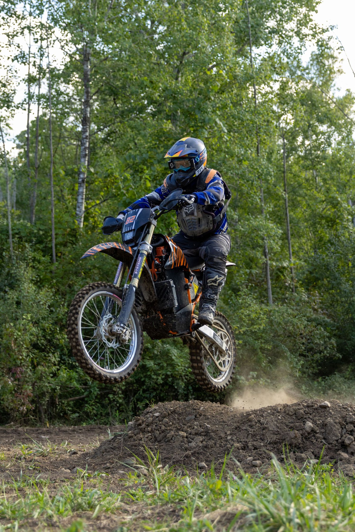 Dirt bike rider catching air while jumping over a dirt mound in a wooded area, spraying dust in dramatic sequence.