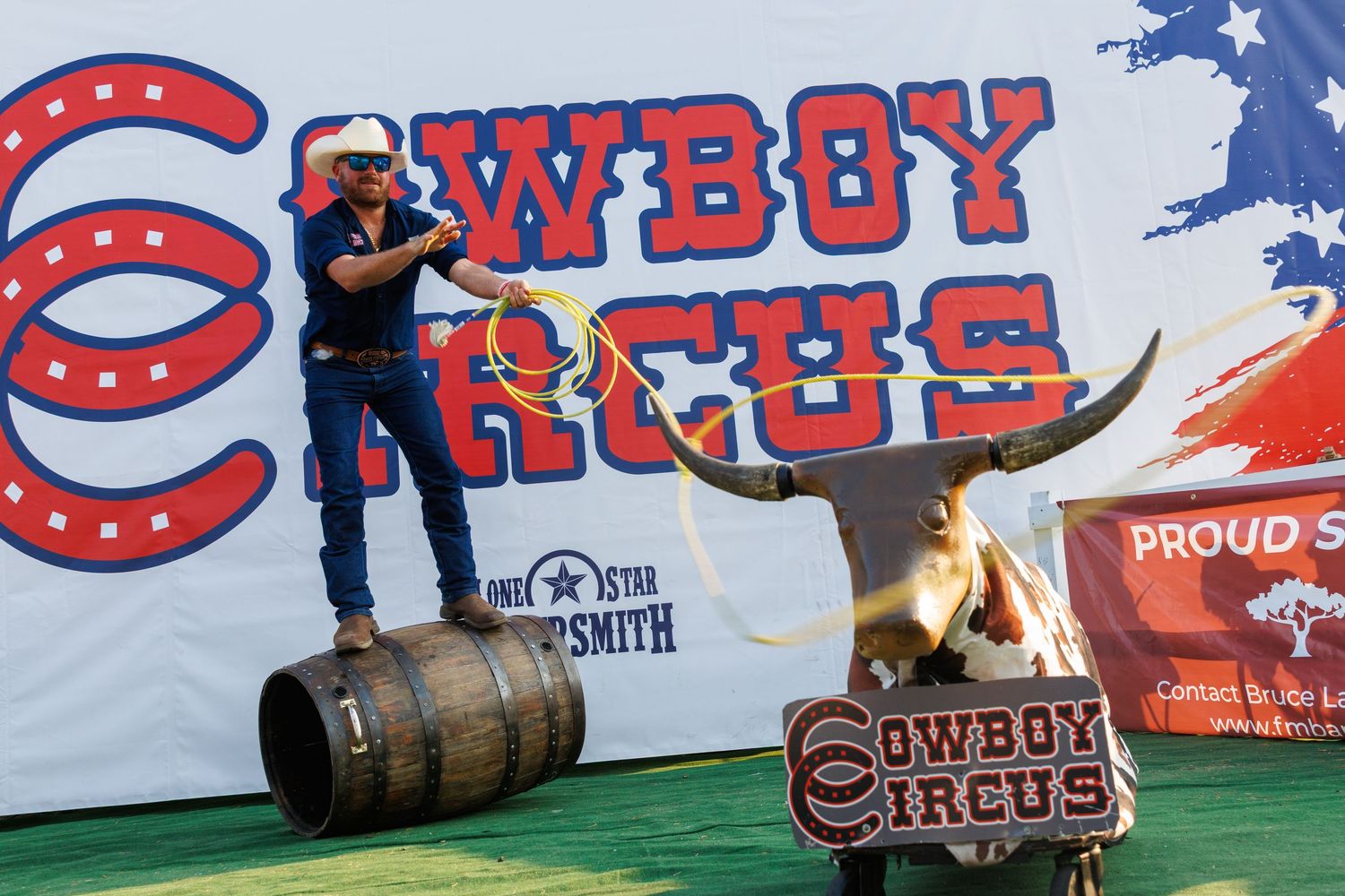 Cowboy performer stands on barrel with lasso at Cowboy Circus event beside golden longhorn bull statue.