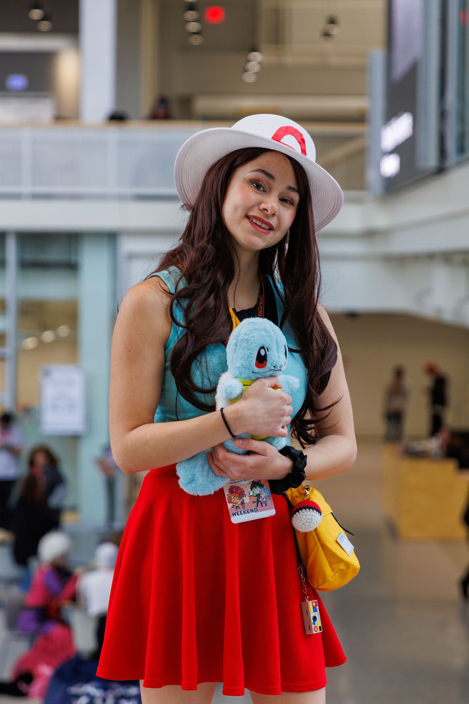 Young woman cosplaying as Pokemon trainer, holding Squirtle plush, wearing red skirt and white hat at convention.