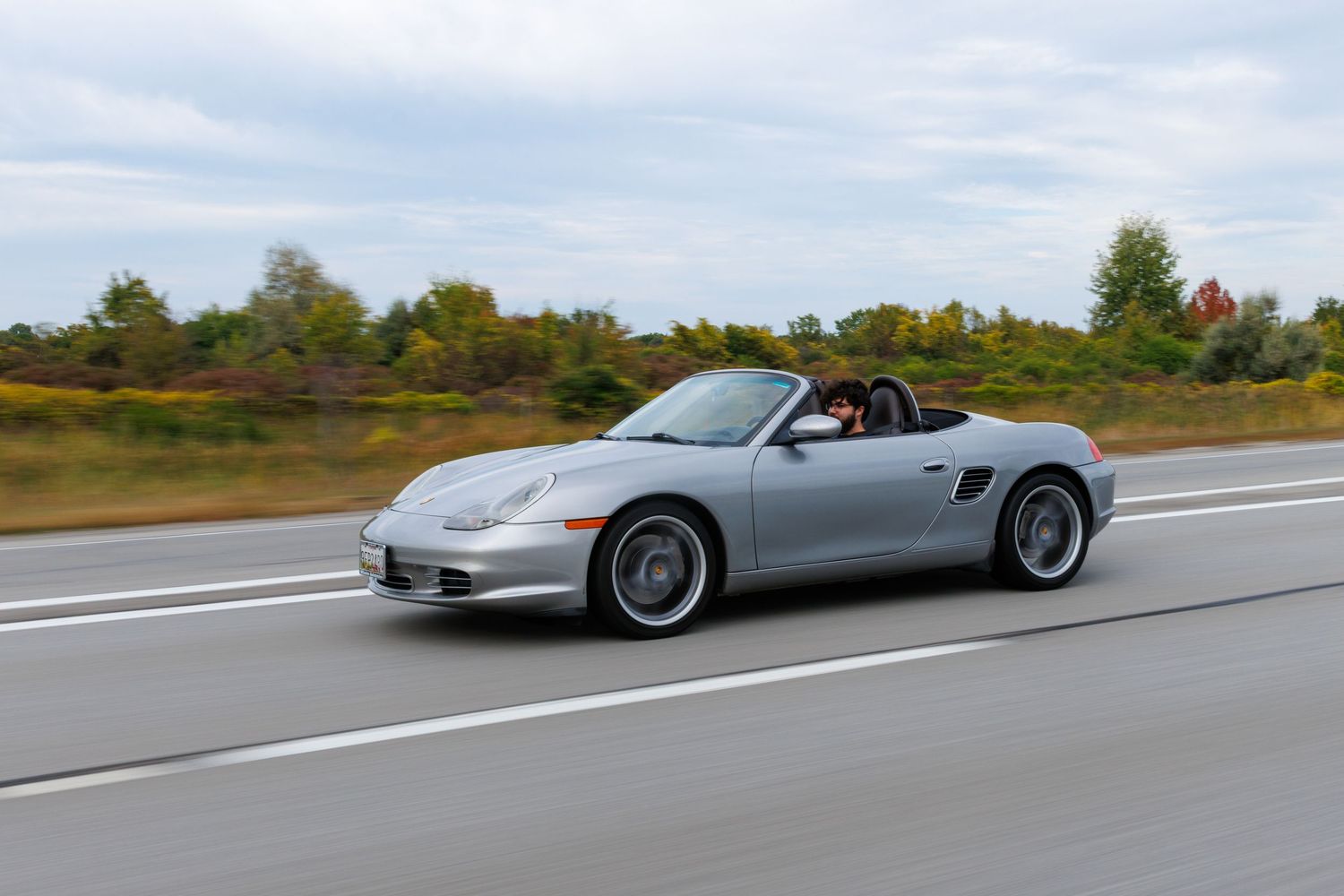 A silver Porsche Boxster convertible sports car speeds along a highway with autumn foliage in the background.