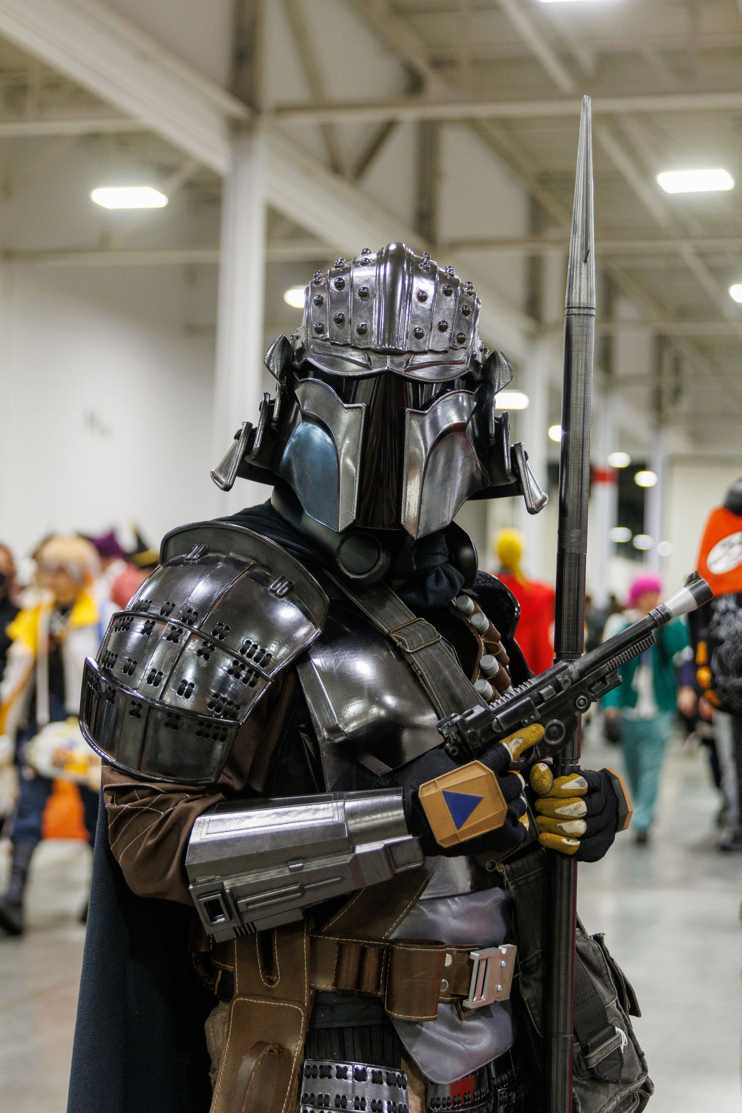 Impressive Mandalorian cosplay featuring detailed silver armor, helmet, cape, and spear at a comic con.