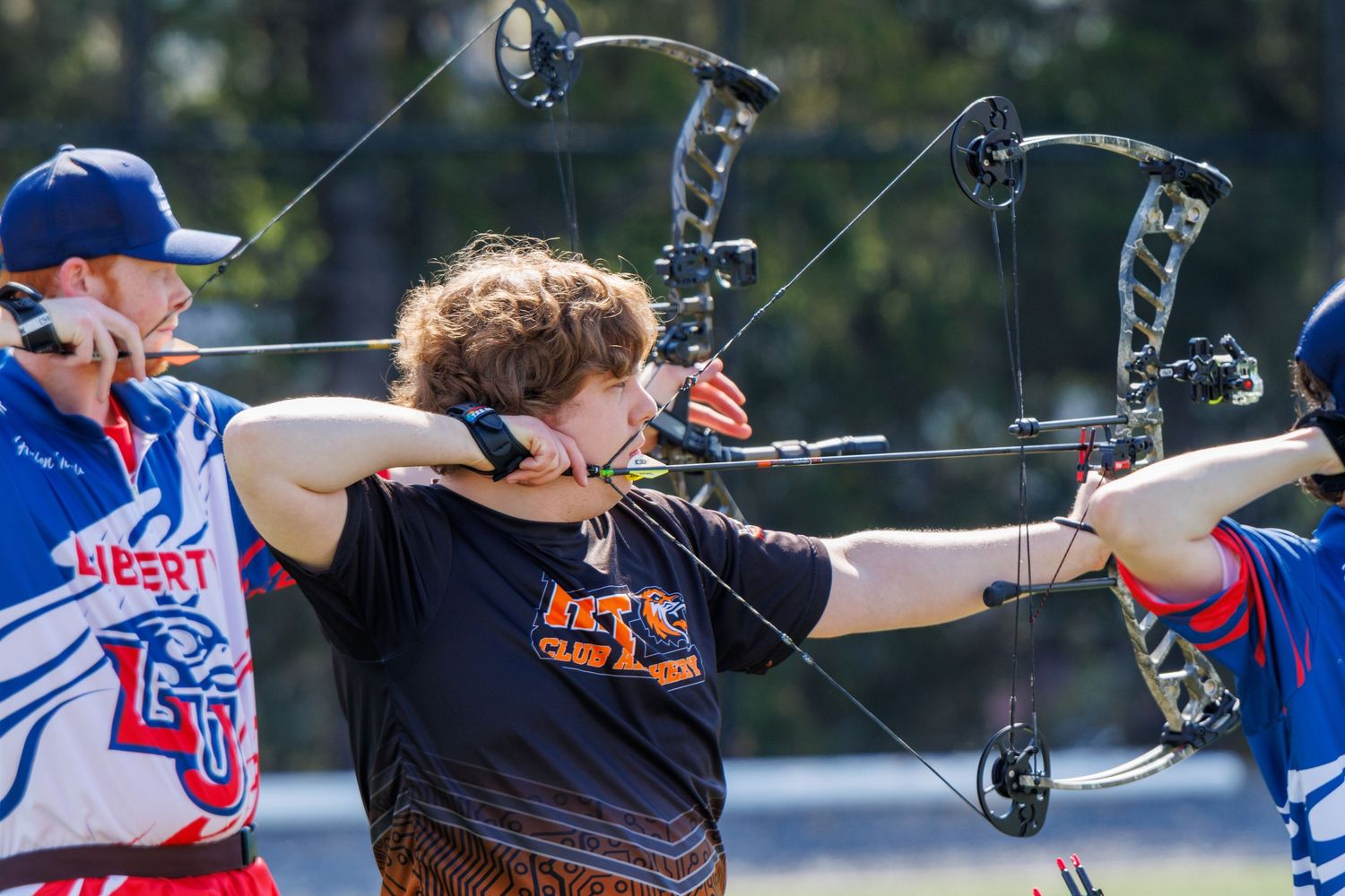 Female archer aims compound bow at target during outdoor archery competition with coach nearby.