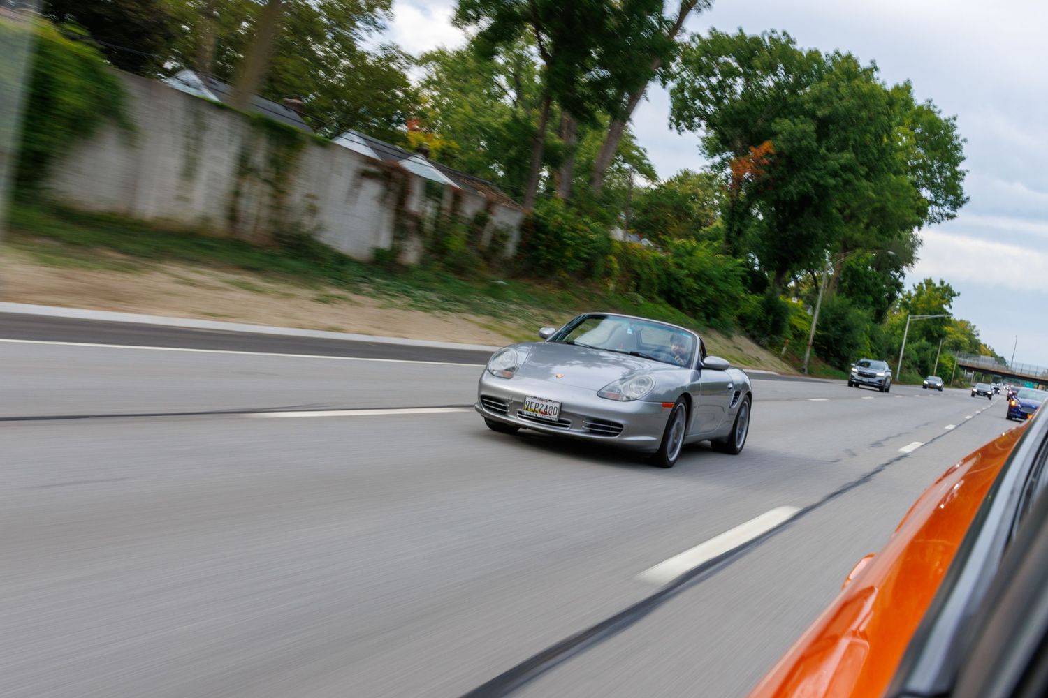 A silver sports car drives past on a curving mountain road with lush green trees in the background.