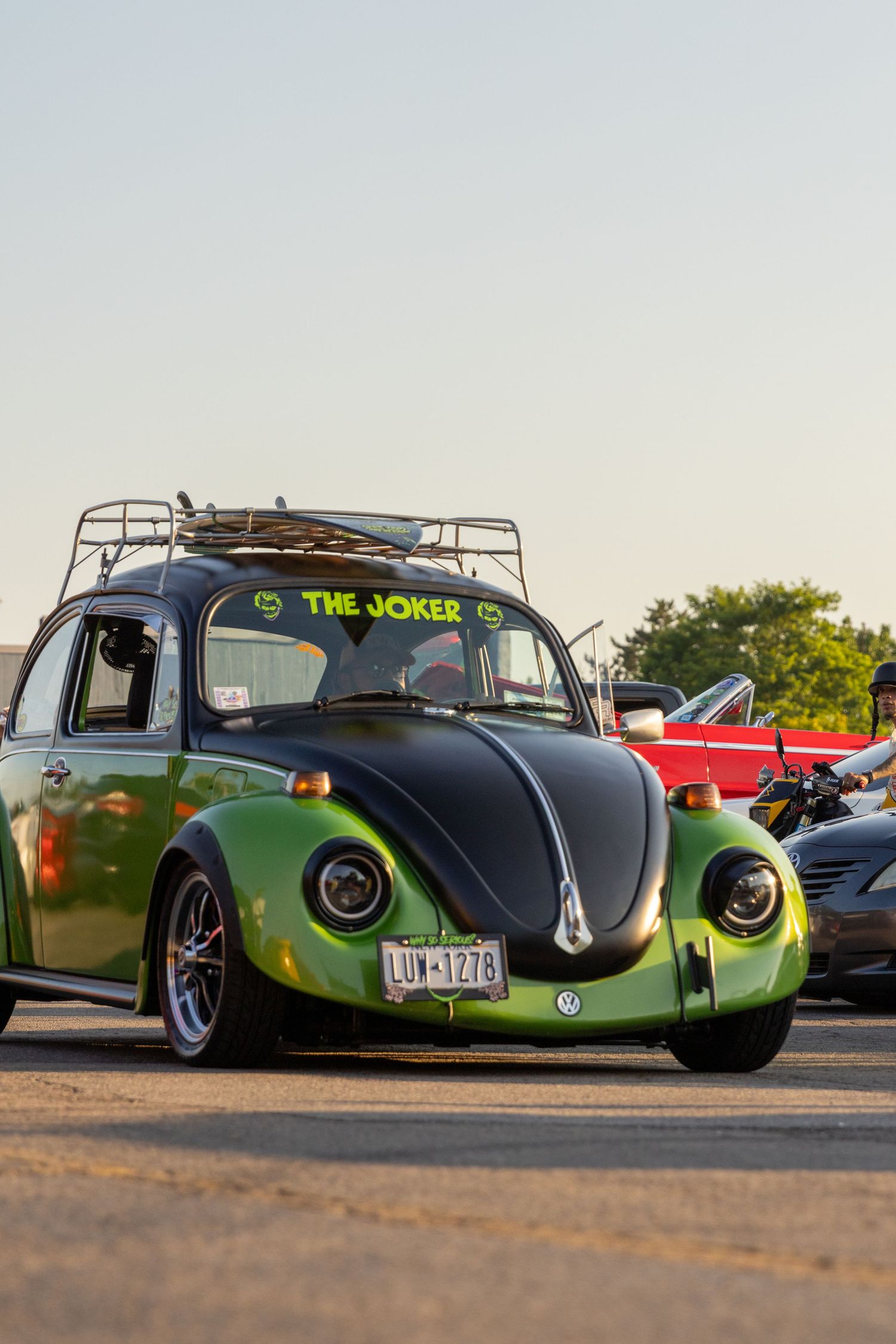 Custom Volkswagen Beetle in green and black paint with 'The Joker' text and roof rack parked at a car show at sunset.