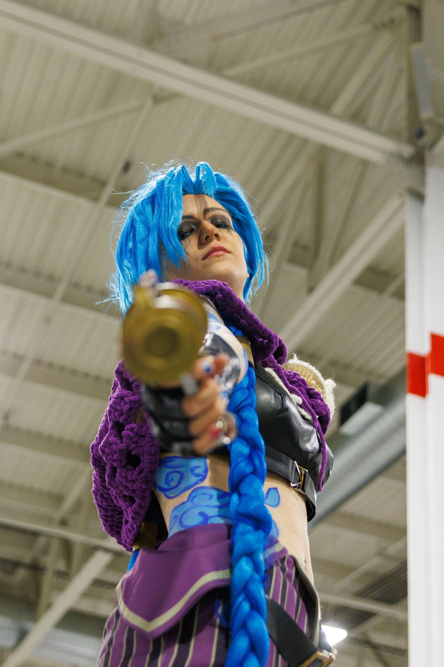 Cosplayer dressed as Jinx from League of Legends with blue hair aims a gold cannon prop at camera.