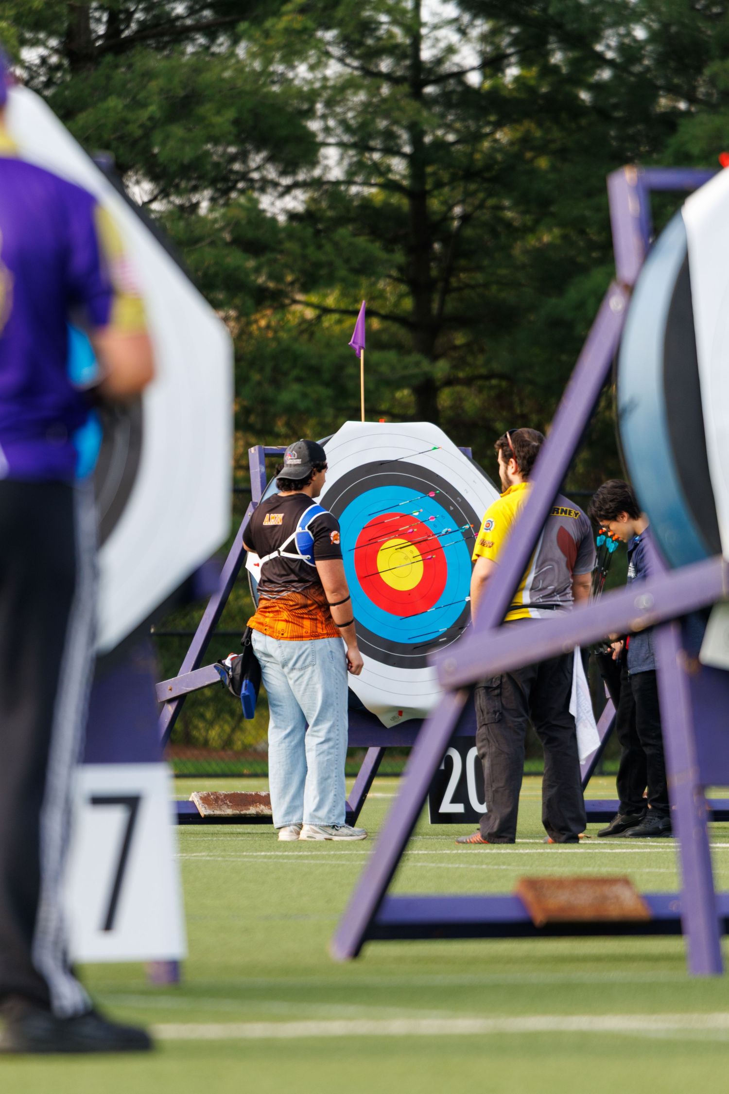 Archers retrieve arrows from colorful target during outdoor archery competition on green field.