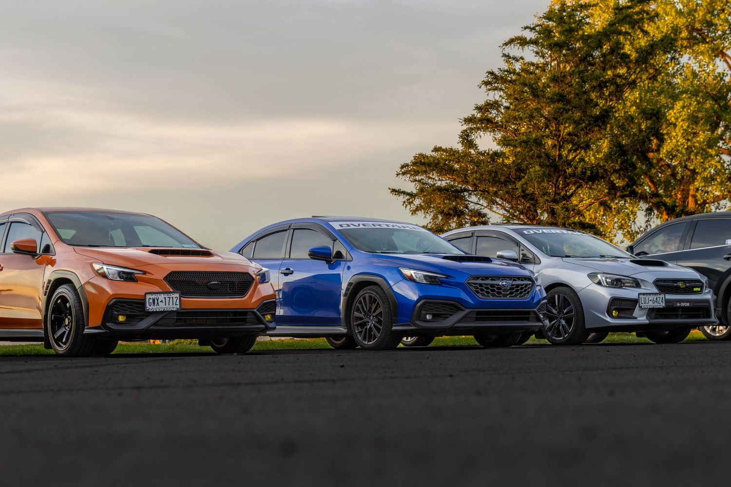 Three Subaru performance cars in copper, blue and silver lined up on display at sunset with autumn trees in background.