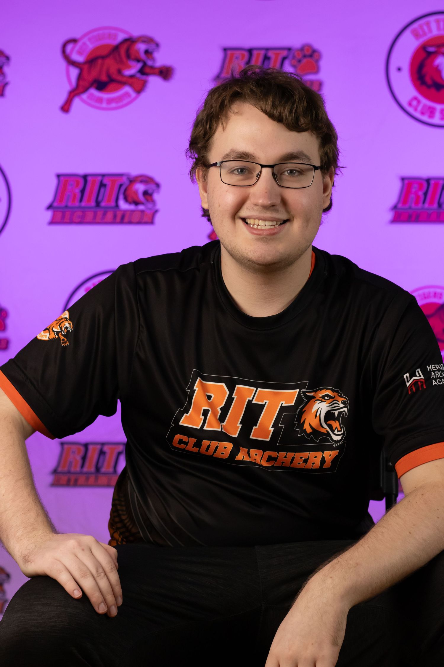 Club hockey player wearing RIT branded gear smiles in front of purple backdrop with tiger logos.