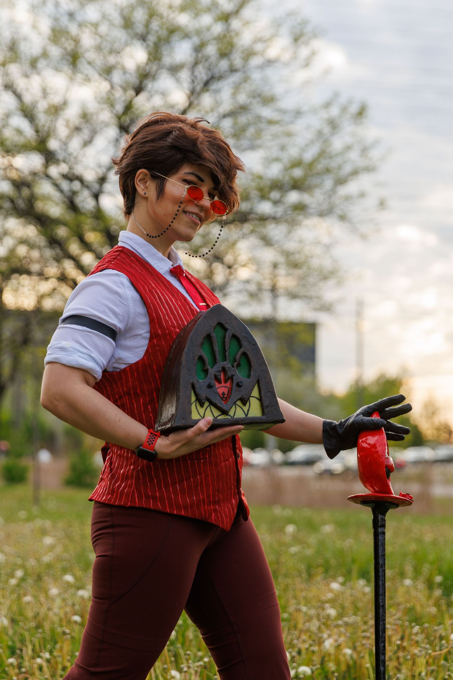 Person cosplaying in a red vest with round glasses holding a vintage radio prop outdoors.