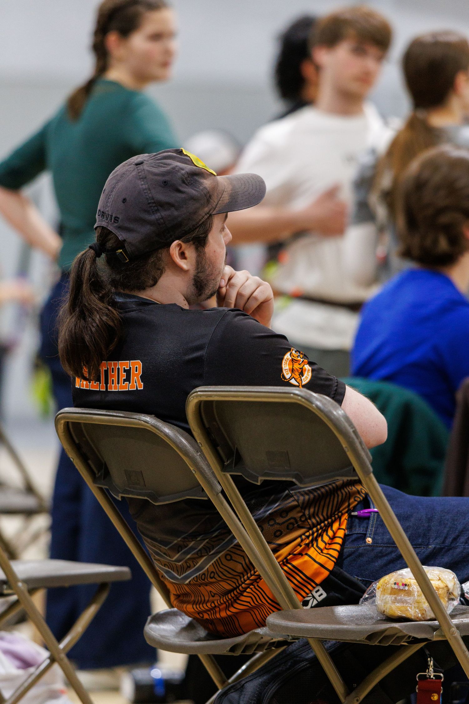 Man with long hair and black shirt sitting in folding chair at an indoor archery tournament watching competitors.