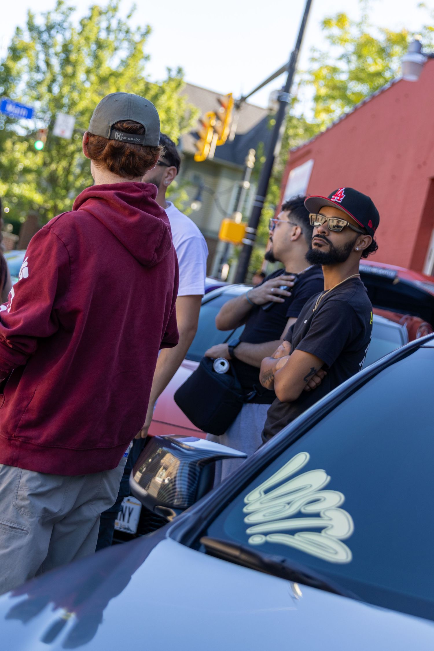 A group gathers around a parked car with a maroon-clad figure visible in the evening light.