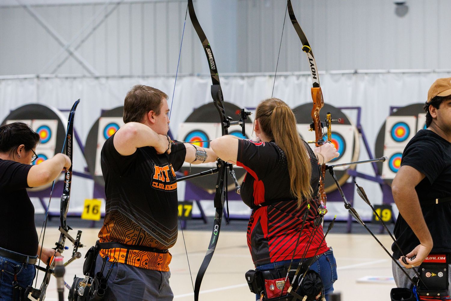 Archers competing indoors at an archery range, drawing bows aimed at colorful target boards in a gymnasium.
