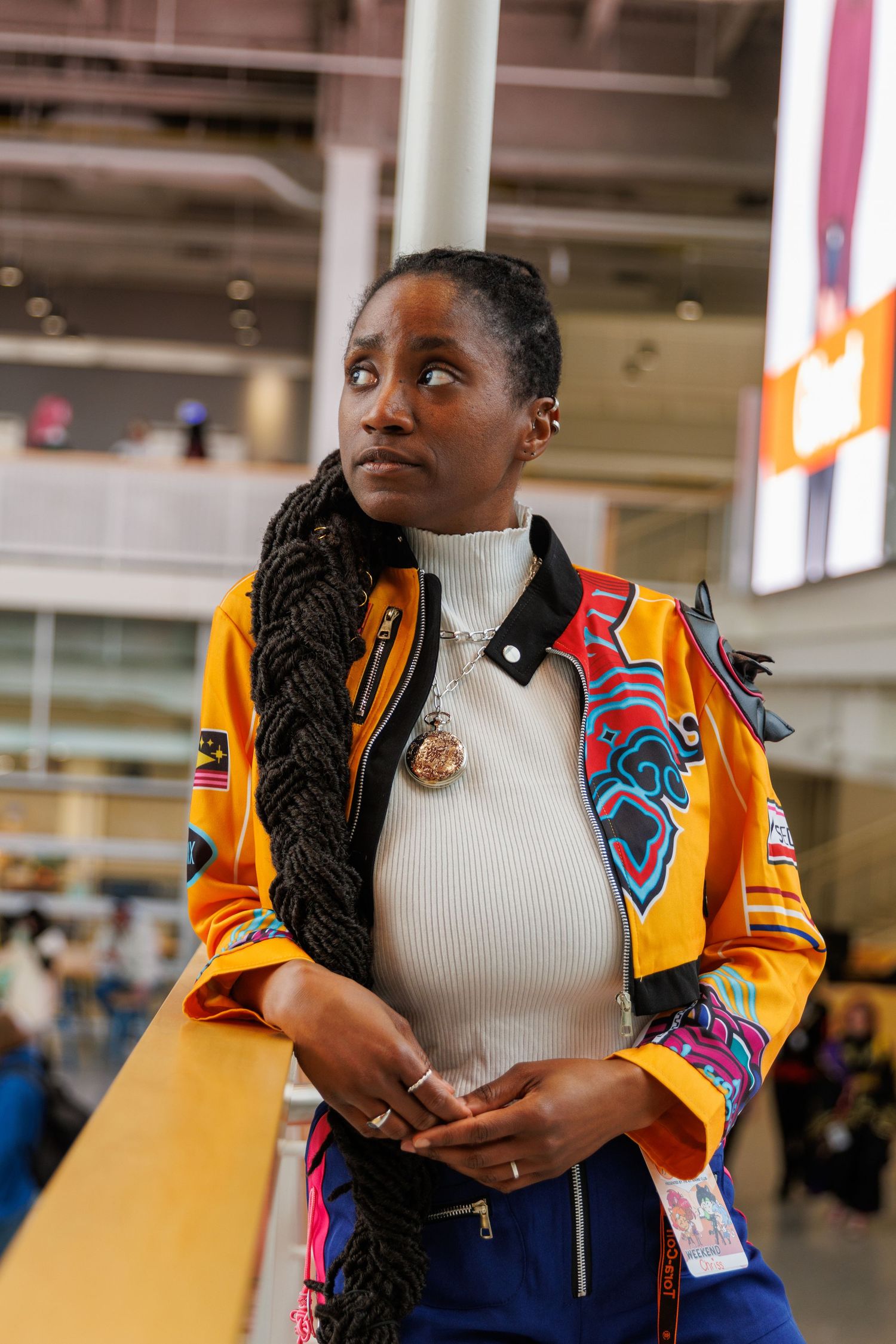 Woman in colorful embroidered orange jacket with braids poses indoors at modern venue.