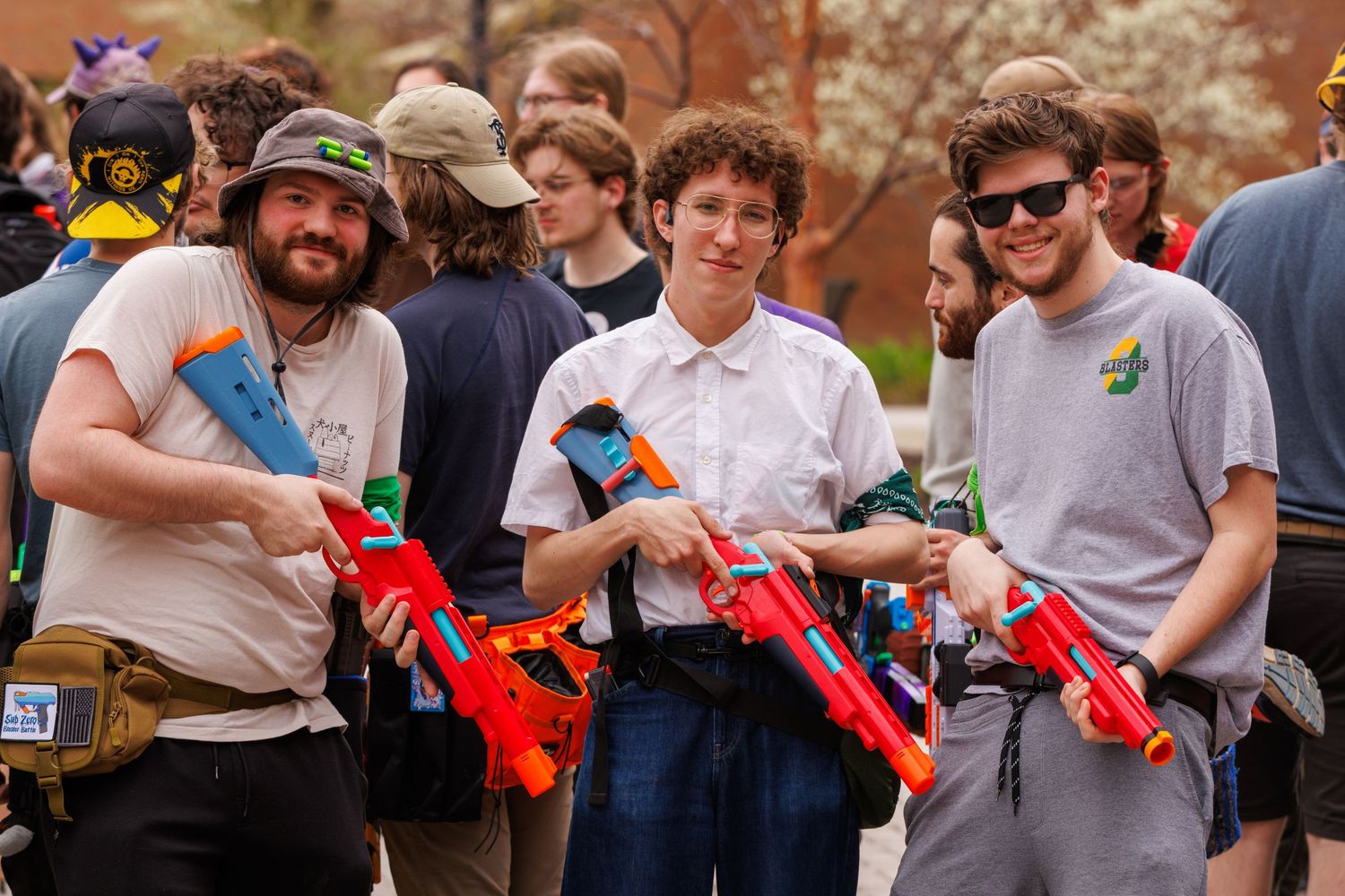 Three young men smiling and holding colorful Nerf guns outdoors at a campus event.