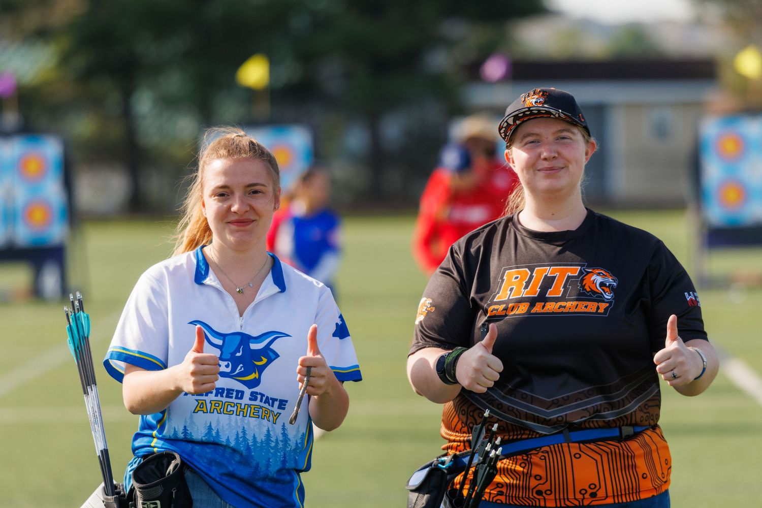Two female archers giving thumbs up at an outdoor archery competition, wearing team uniforms.