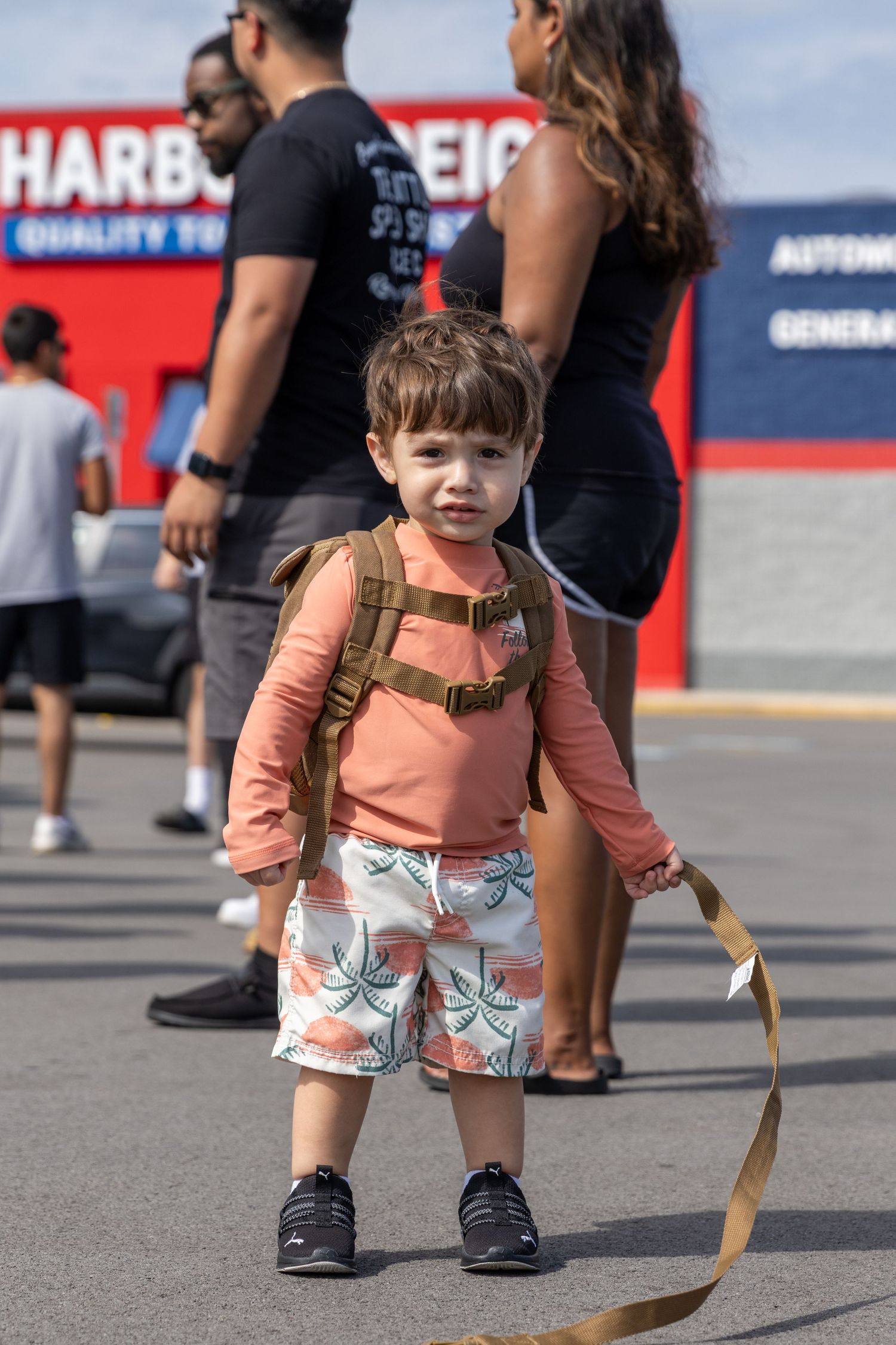 Young child wearing a peach colored shirt and printed shorts stands on a parking lot surface near a storefront.