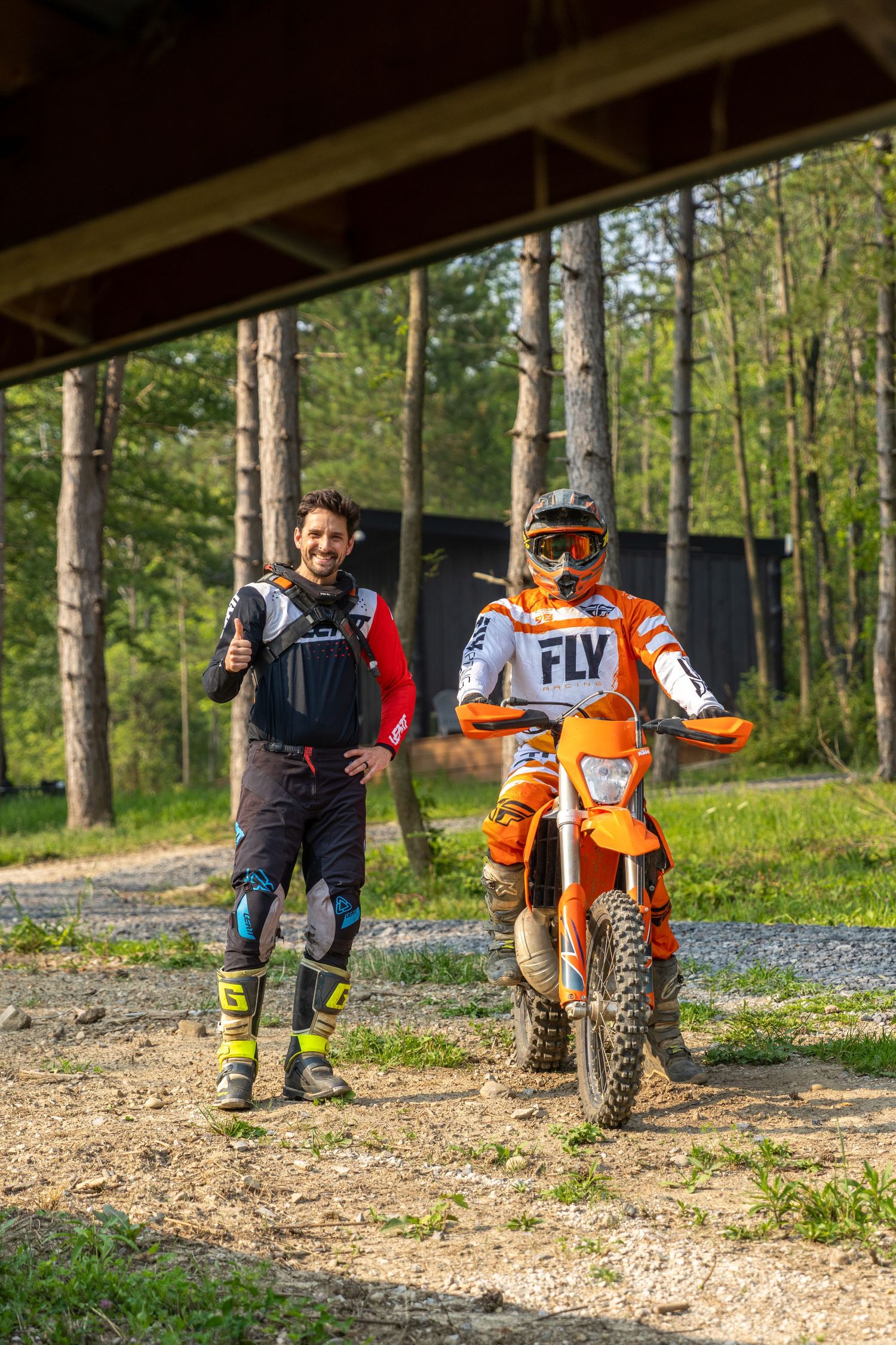 Dirt bike riders taking a break on a wooded trail with their orange and black motocross bikes on a sunny day.