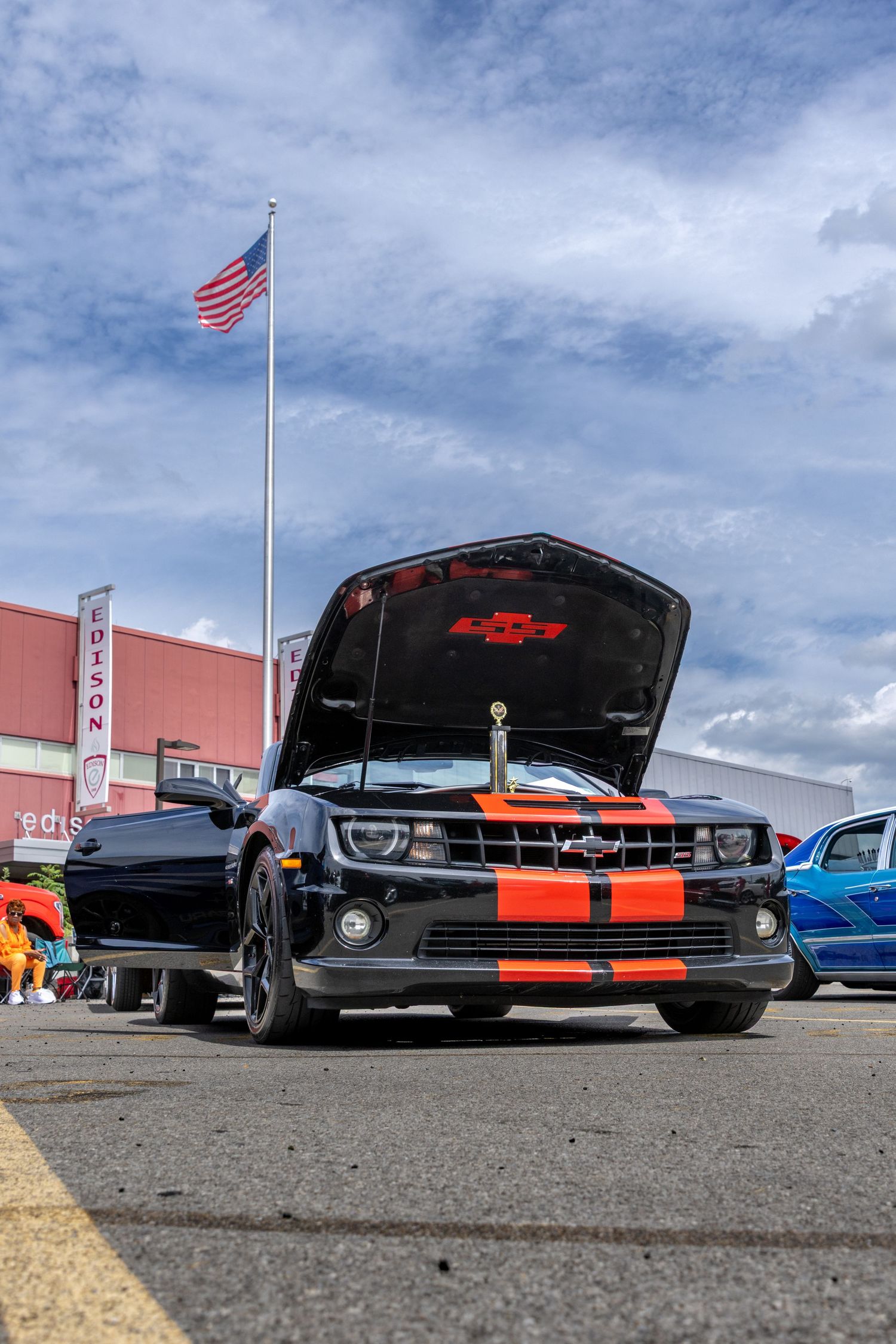 Classic cars with open hoods on display in a parking lot under cloudy skies with an American flag waving.