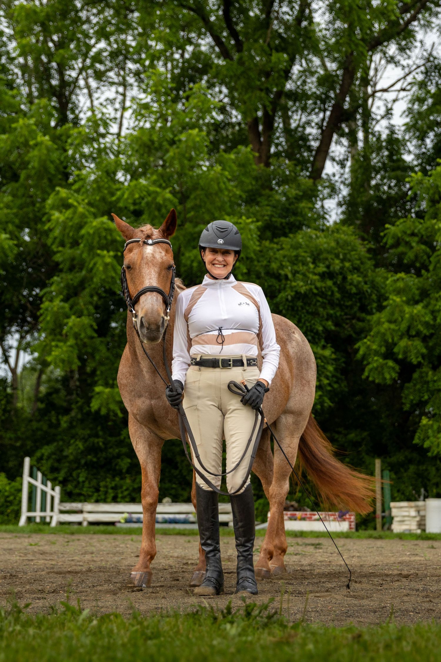 An equestrian poses with a chestnut horse in full riding attire on a dirt arena.