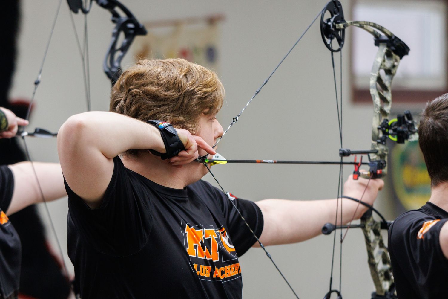 Young archer in black team shirt draws compound bow during indoor archery practice session.