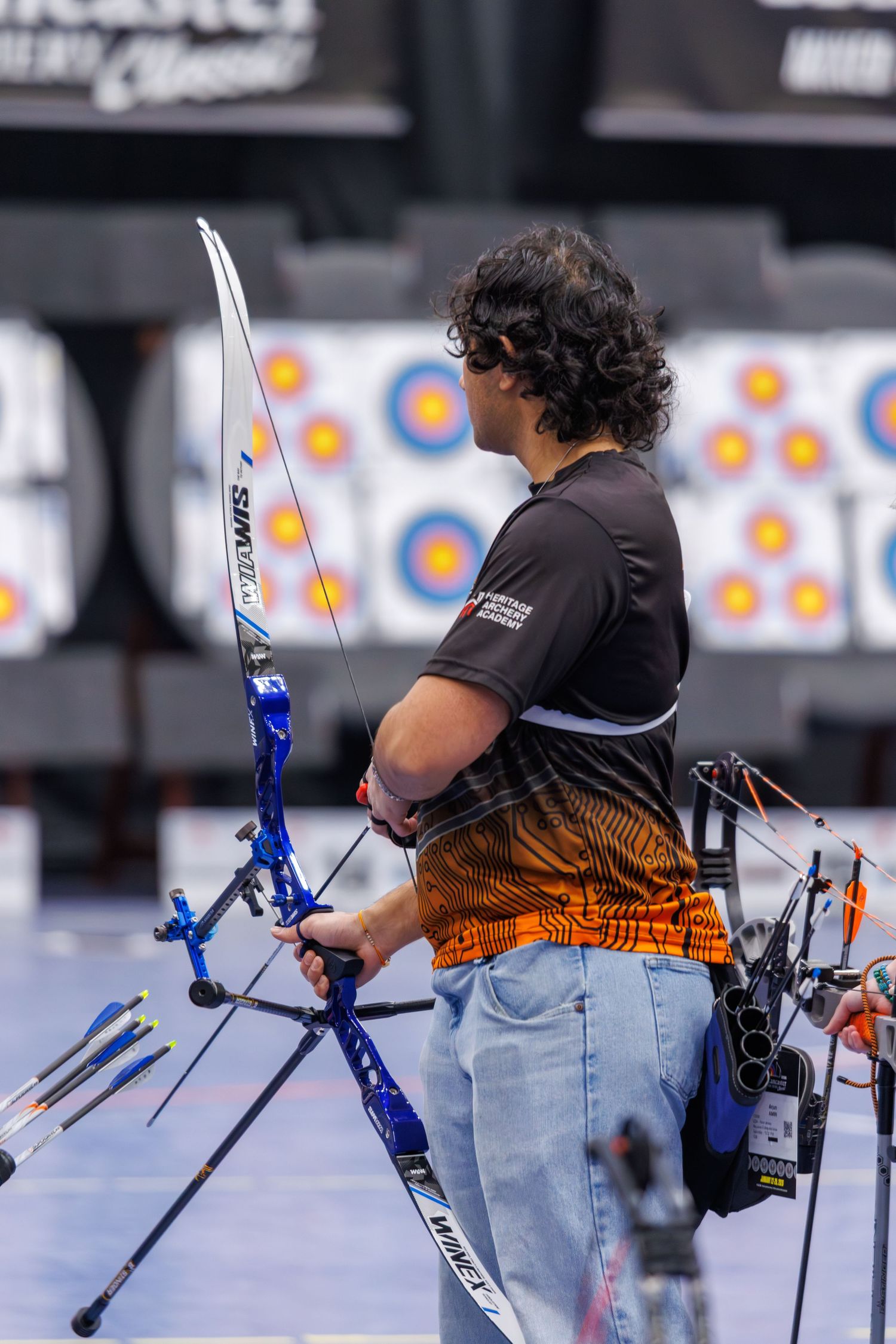 Archer competing indoors at a classic tournament, aiming a blue recurve bow at colorful targets.